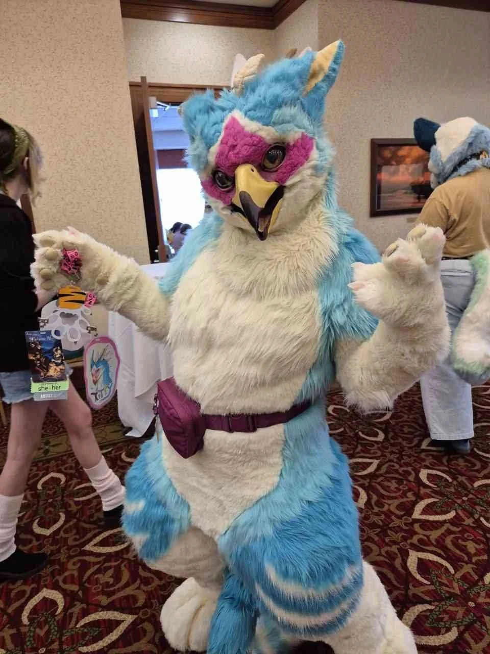 Person dressed in a colorful, furry blue and white gryphon costume with pink accents, posing with arms bent at the elbows in a crowded indoor event.