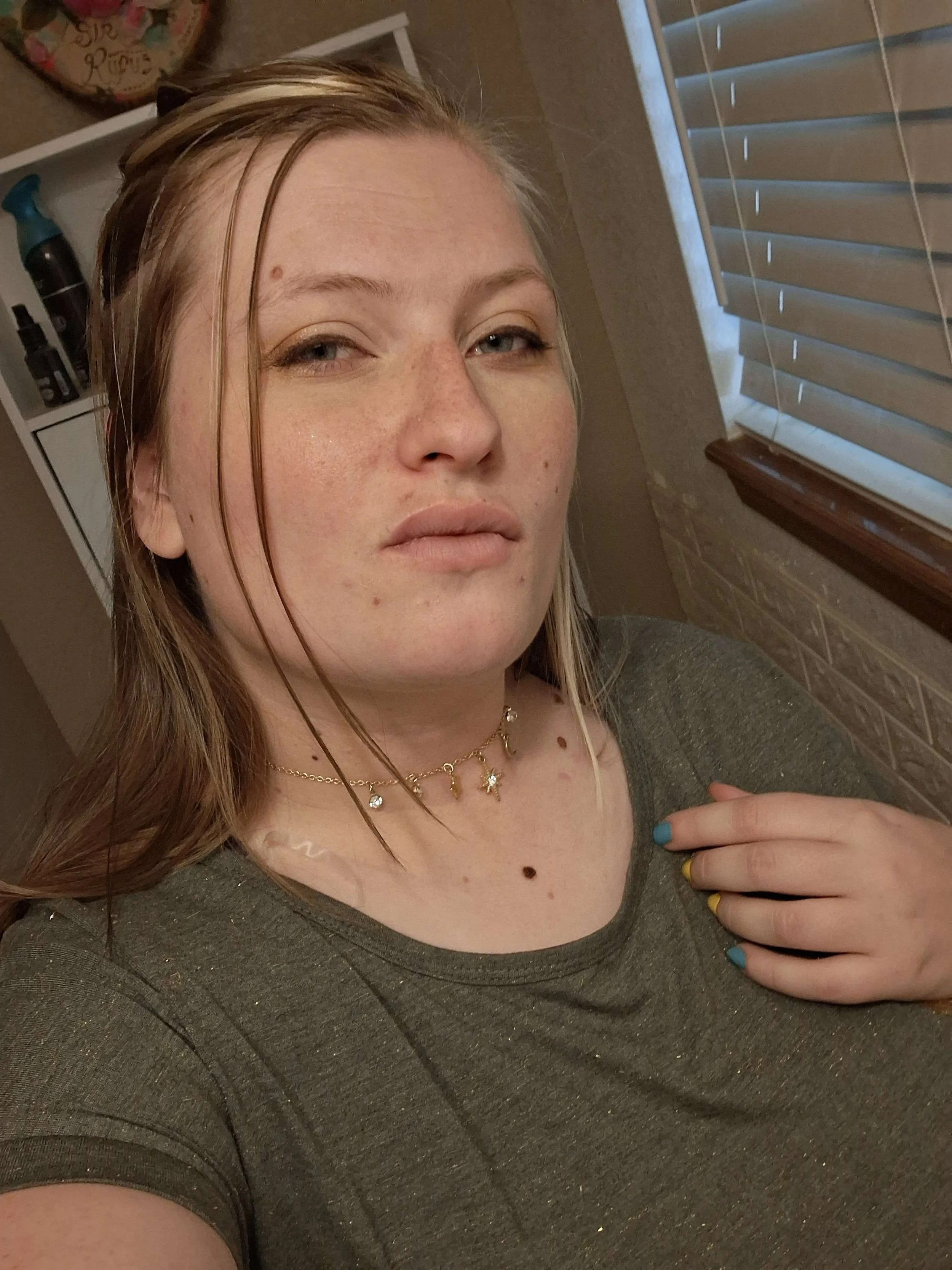 A young woman with long, lightly damp blonde hair, wearing a green t-shirt, gold necklaces, and nail polish with blue and yellow colors, taking a selfie in a room with a window and a shelf in the background.