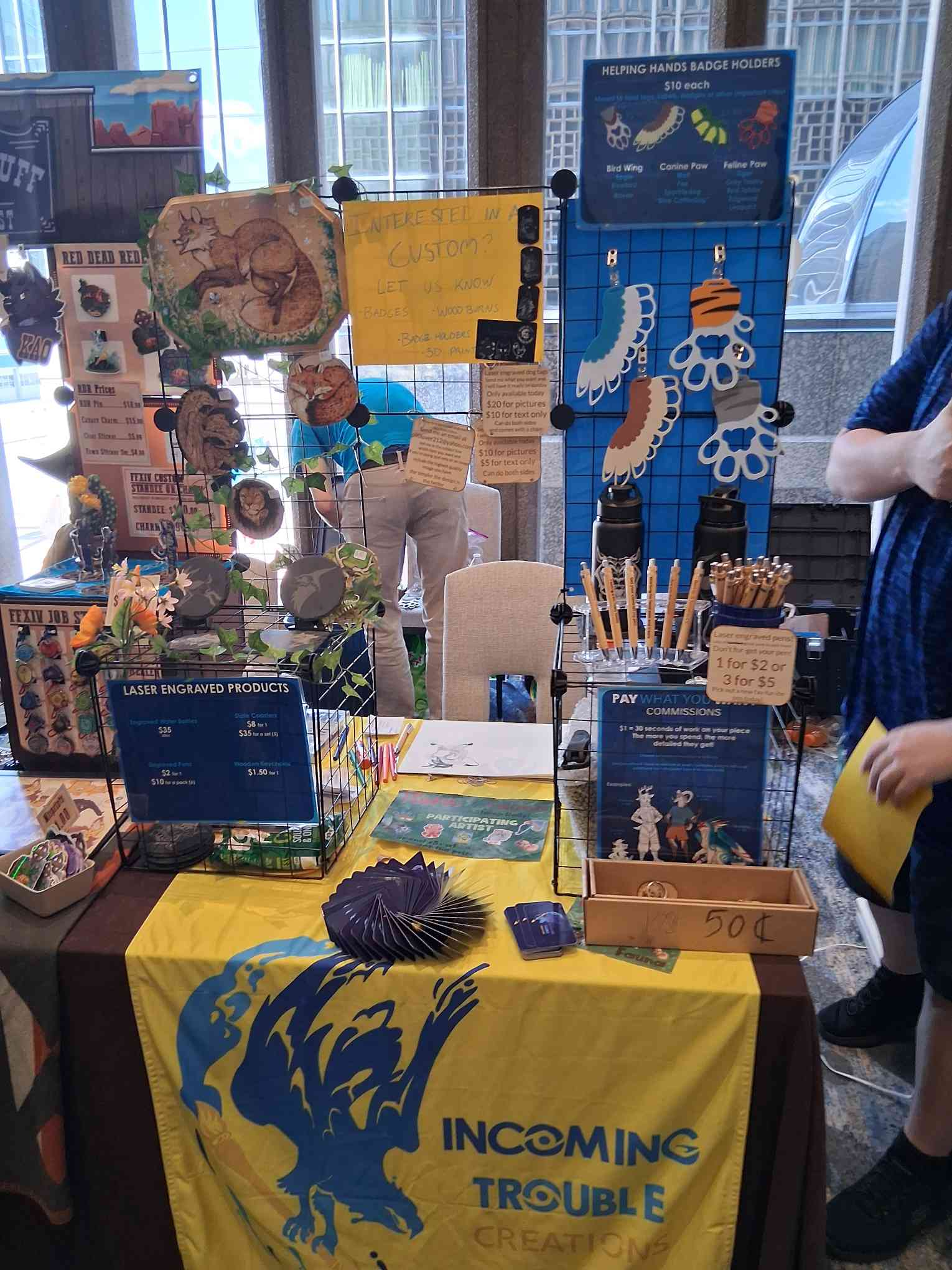 Table with various handcrafted items for sale including engraved products, badges, and bird wings for badge holders. Signage indicates prices and custom options, with a yellow banner featuring a blue mythical creature and the words "Incoming Trouble Creations." In the background, a blue display board showcases more badges and signs advertising badge holder customization services.