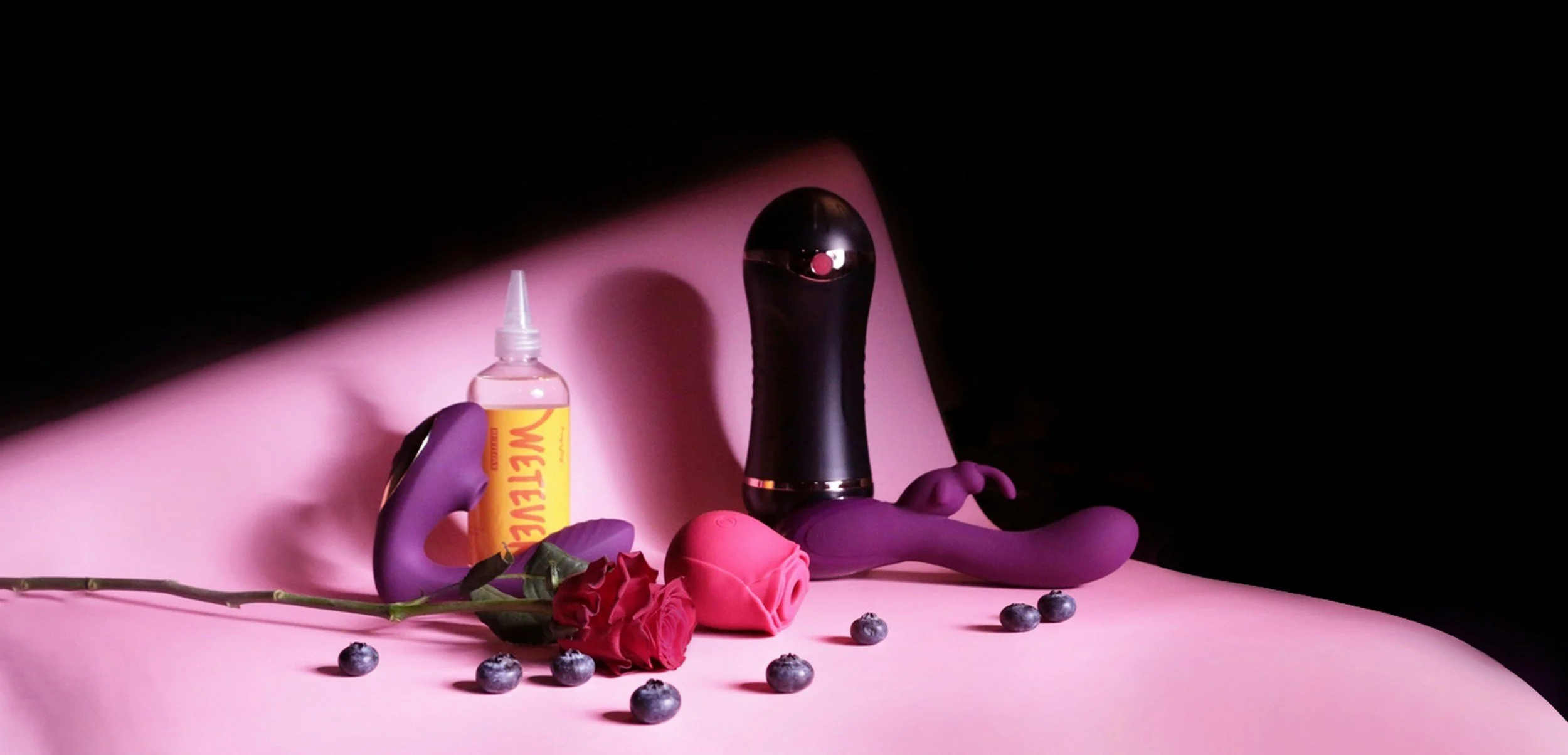 A still life with a pink background, featuring adult toys, a rose, blueberries, and a bottle of lubricant.