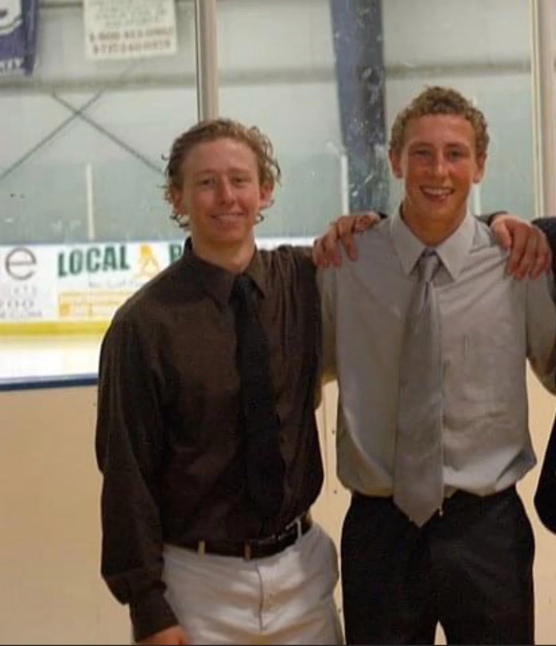 Two young men in dress shirts and ties standing on an ice hockey rink, smiling and posing with arms around each other's shoulders.