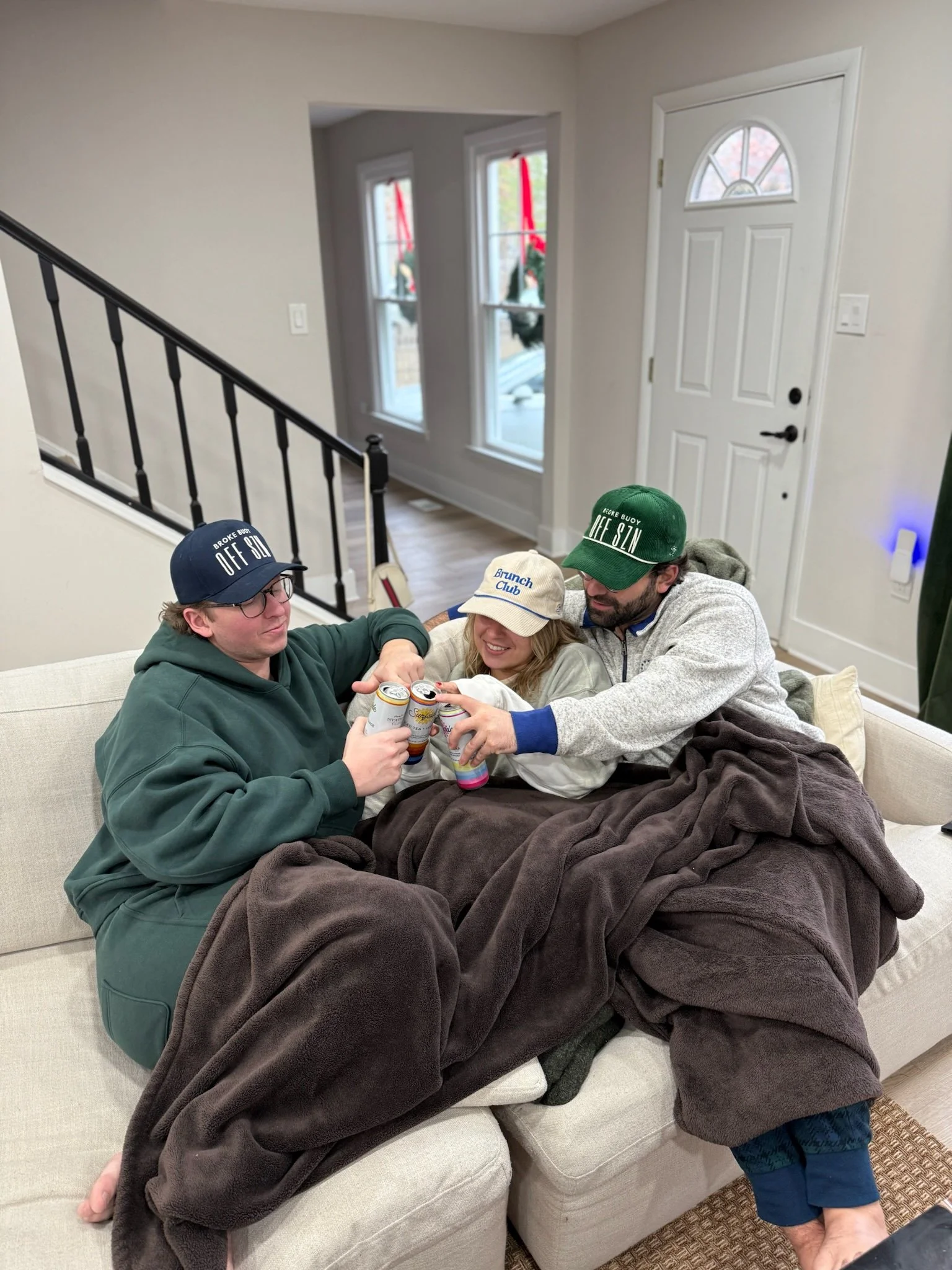 Three friends sitting on a beige couch, wrapped in a brown blanket, toasting with cans of soda.