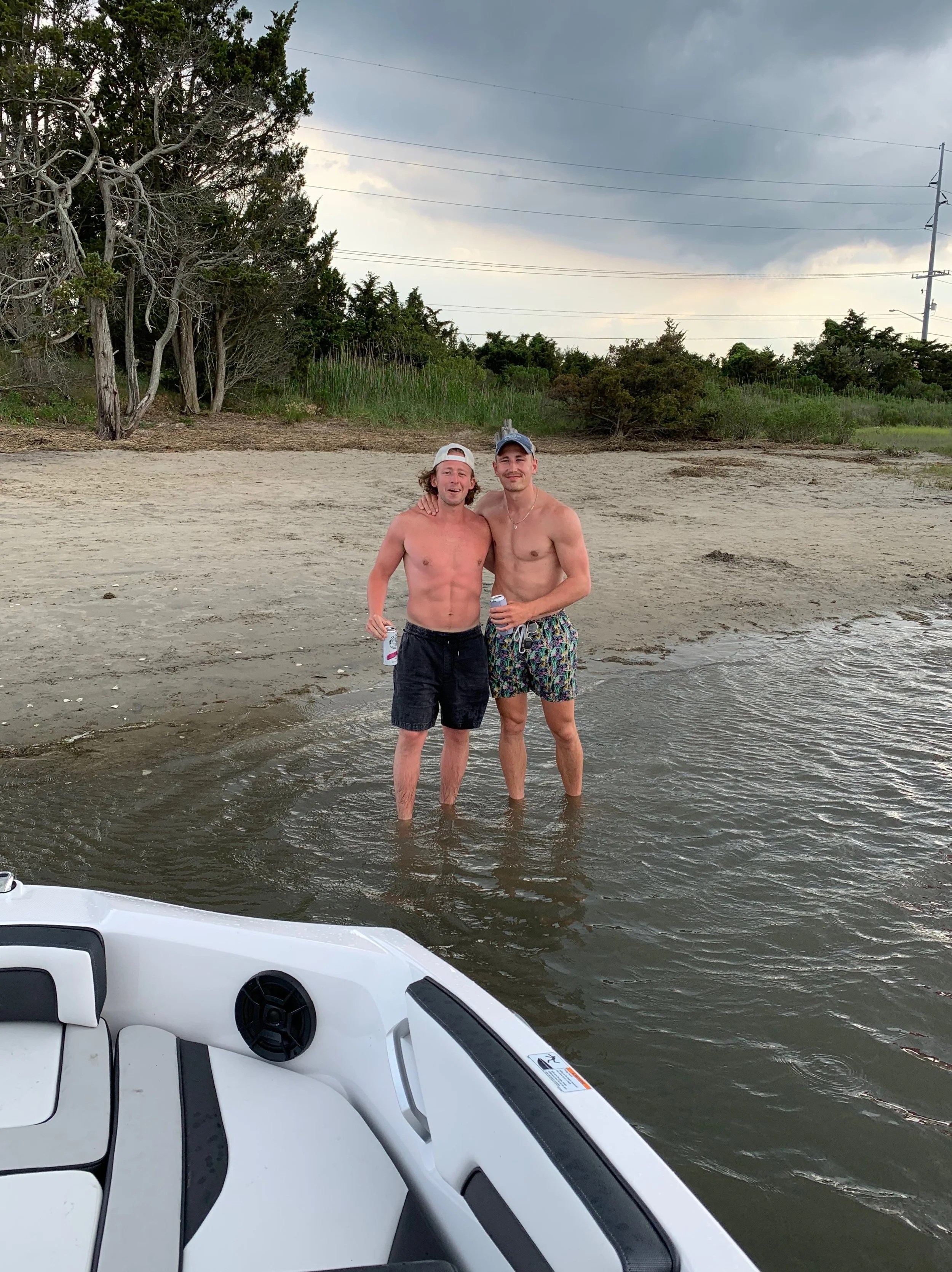 Two young men standing in shallow water at the beach, smiling, with one wearing a baseball cap and the other with long hair, both shirtless, holding drinks. The background shows trees, bushes, and a cloudy sky.