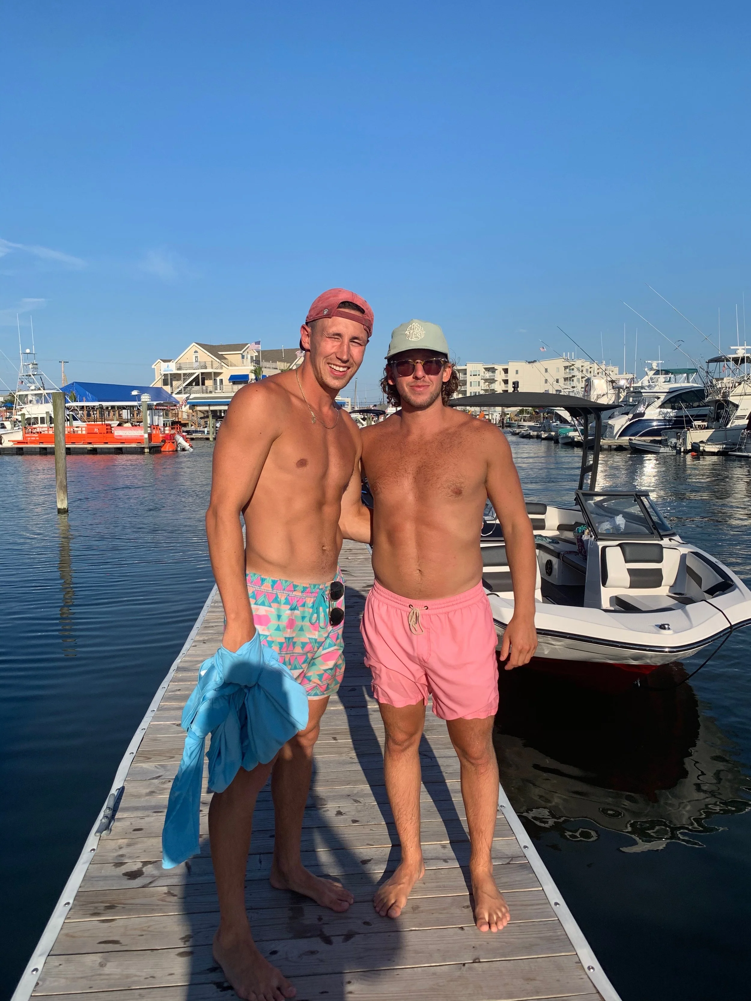 Two shirtless young men standing on a wooden dock by boats in a marina, smiling at the camera. One is holding a blue shirt. They are wearing colorful swim shorts, with the man on the left in bright patterned shorts and the man on the right in pink sh