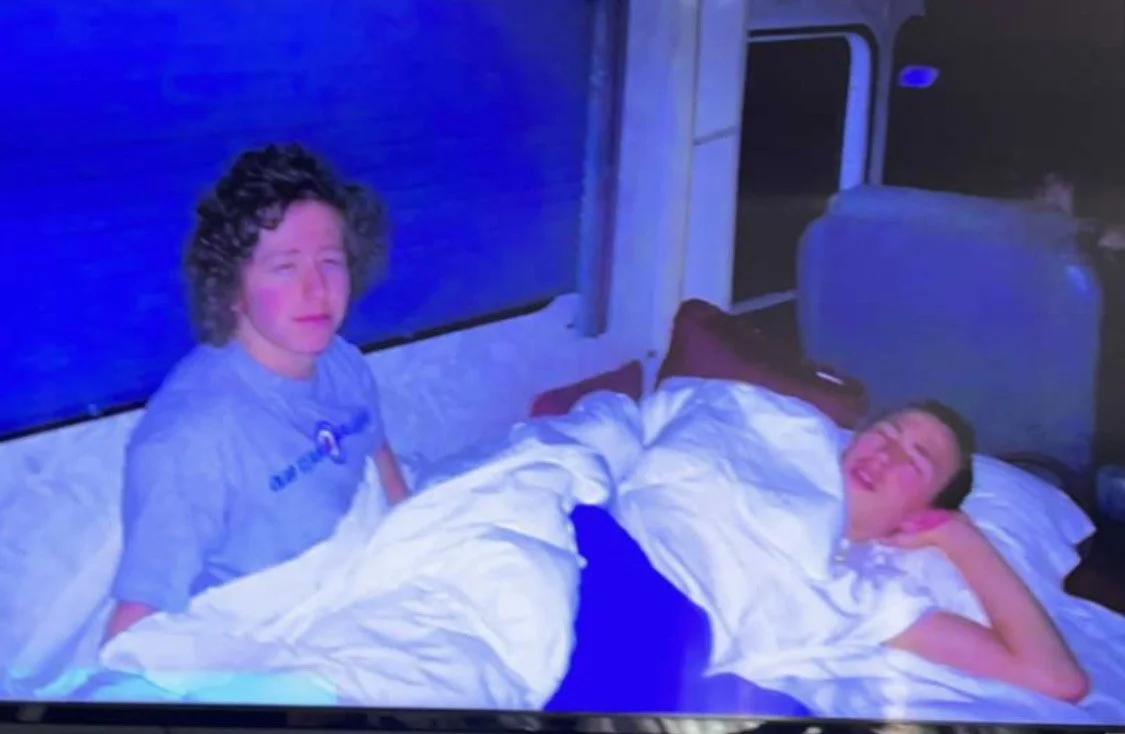 Two young boys in bed, one sitting upright and looking at the camera with a curious expression, the other lying down and smiling, in a bedroom with blue lighting.