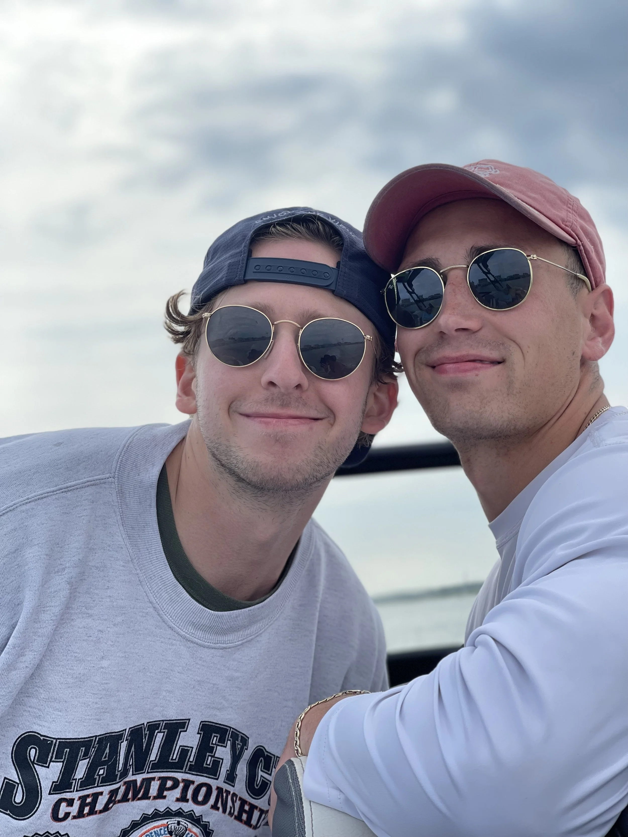 Two young men smiling together, wearing sunglasses and casual clothing, outdoors under a cloudy sky.