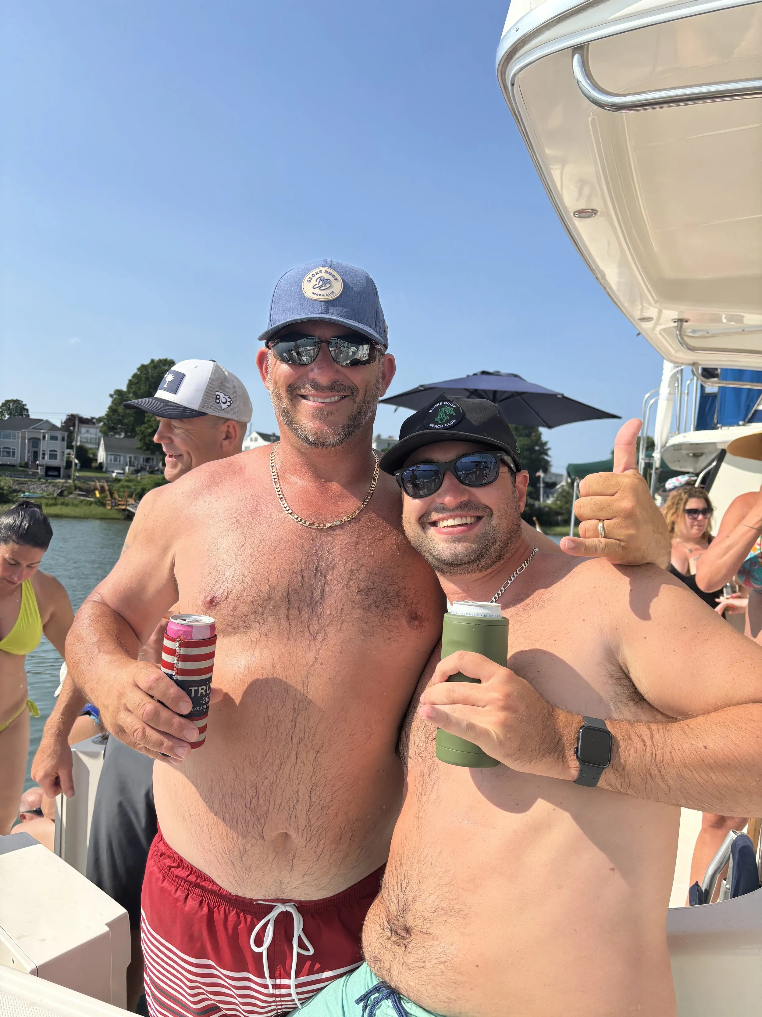 Two men are celebrating outdoors on a boat, both shirtless and wearing sunglasses, holding drinks, smiling and showing friendly gestures. Several other people are visible in the background near water and boats.