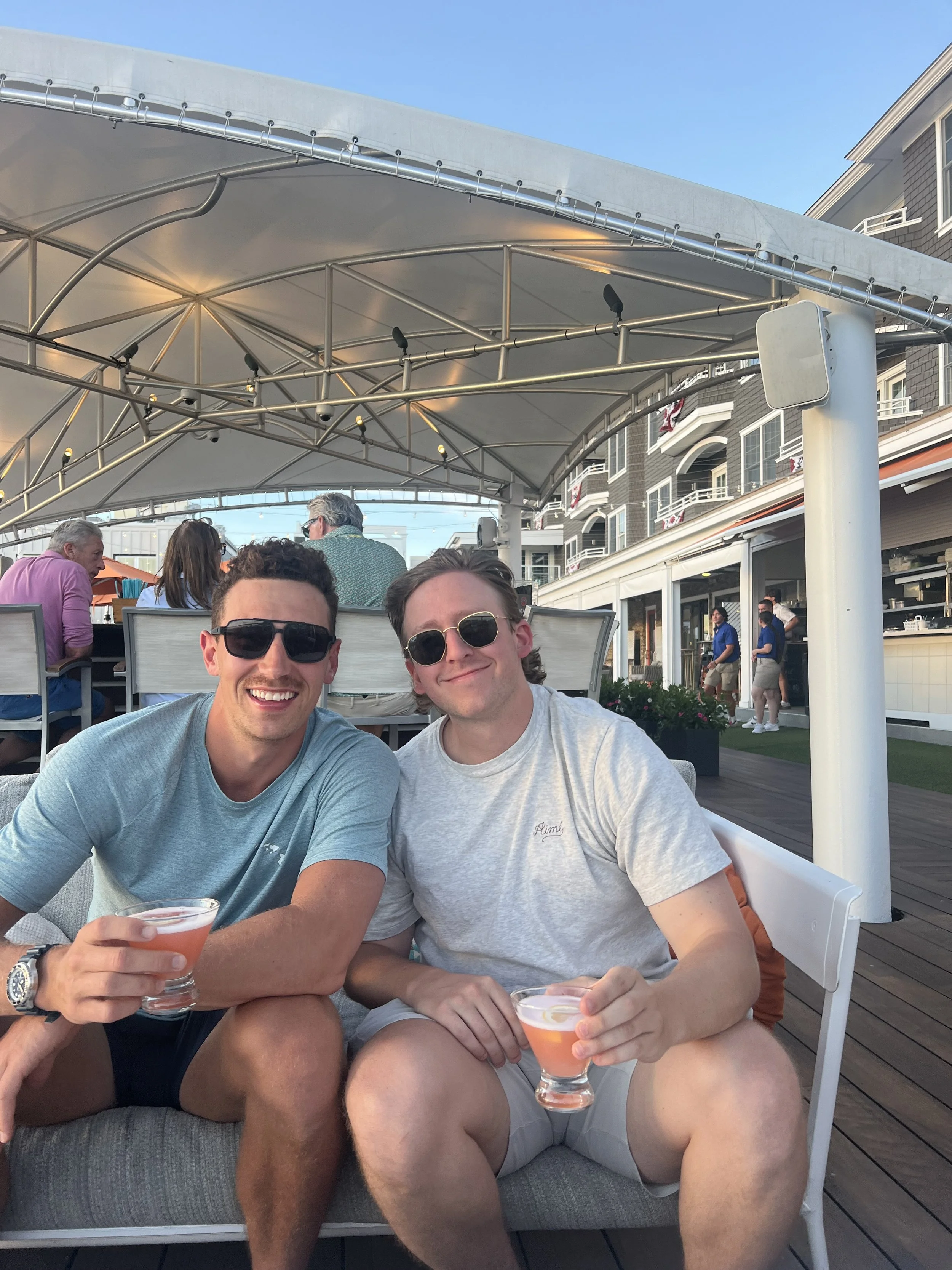Two young men sitting on a terrace, smiling, holding drinks, with a bar and other patrons in the background under a white canopy.