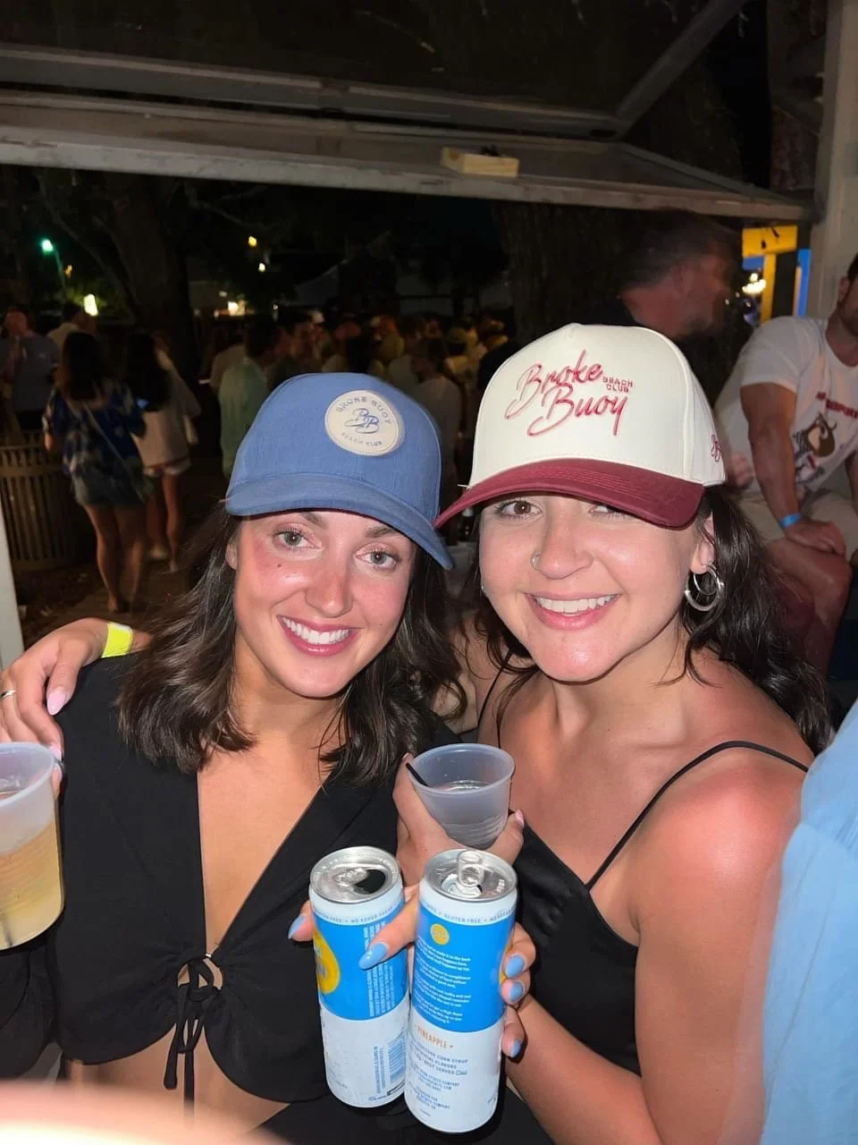 Two women smiling at a beach bar, wearing hats, holding cans of bubbly, and drinks, in a lively outdoor setting at night.