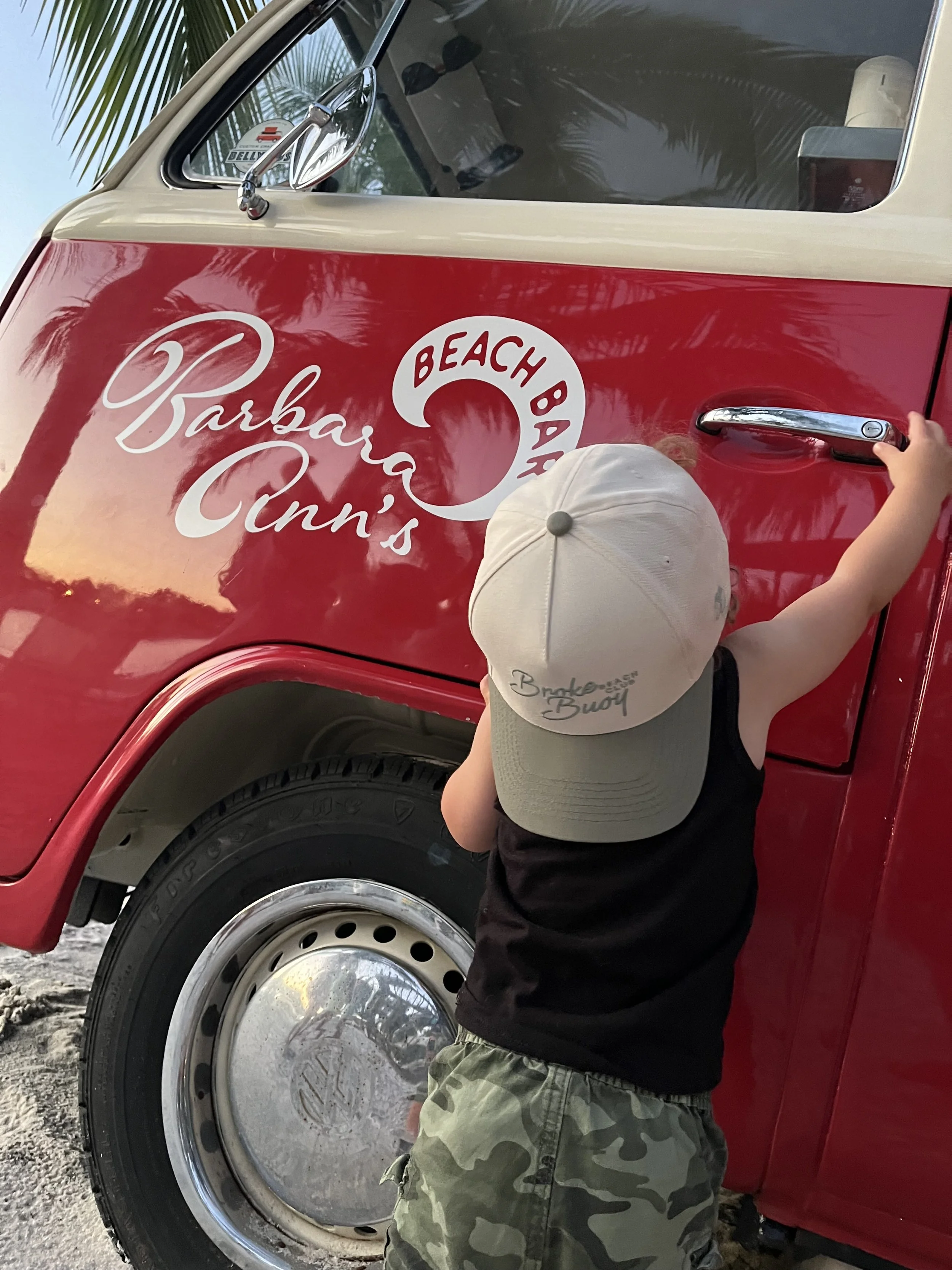 A young child in camouflage shorts and a beige hat stands next to a vintage red and white Volkswagen van with the words 'Barbara Ann's Beach Bar' written on it. The child is reaching for the door handle.