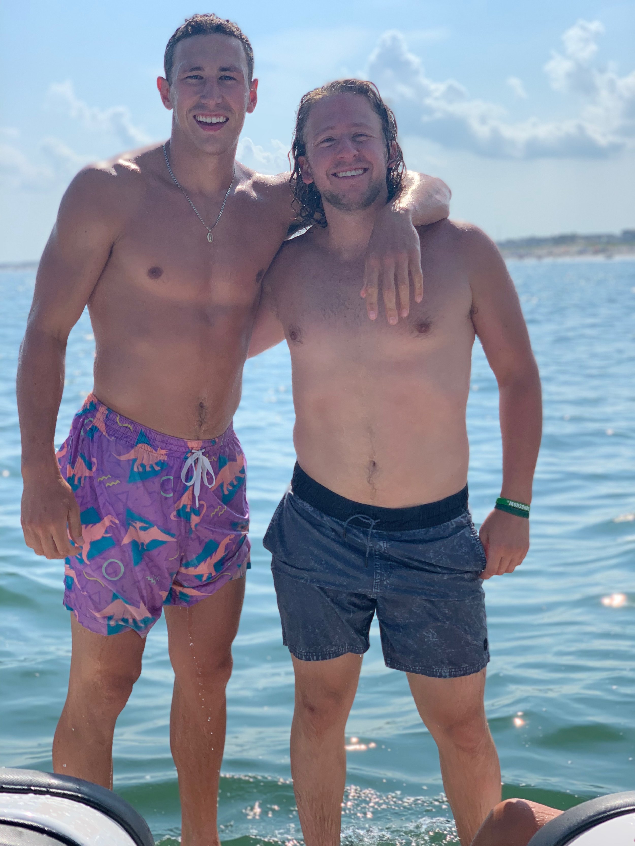 Two shirtless men standing in water with arms around each other's shoulders, smiling at the camera under a partly cloudy sky.
