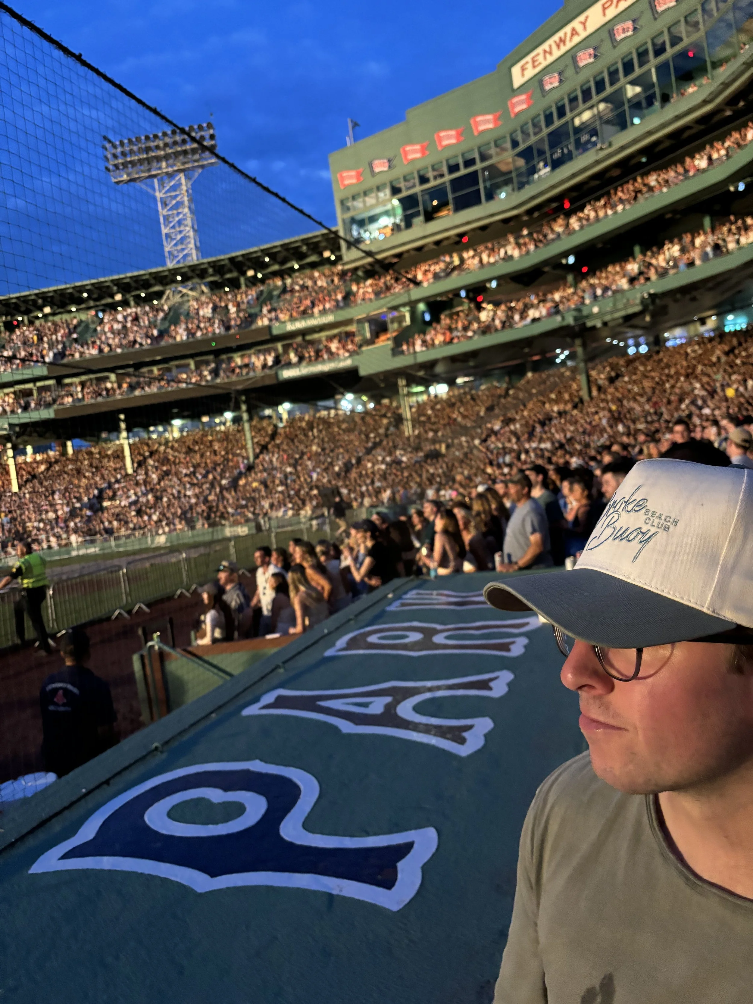 A baseball stadium packed with fans, with a man in glasses and a white cap with blue brim and text, looking down, in the foreground. The stadium is filled with spectators during an evening event.