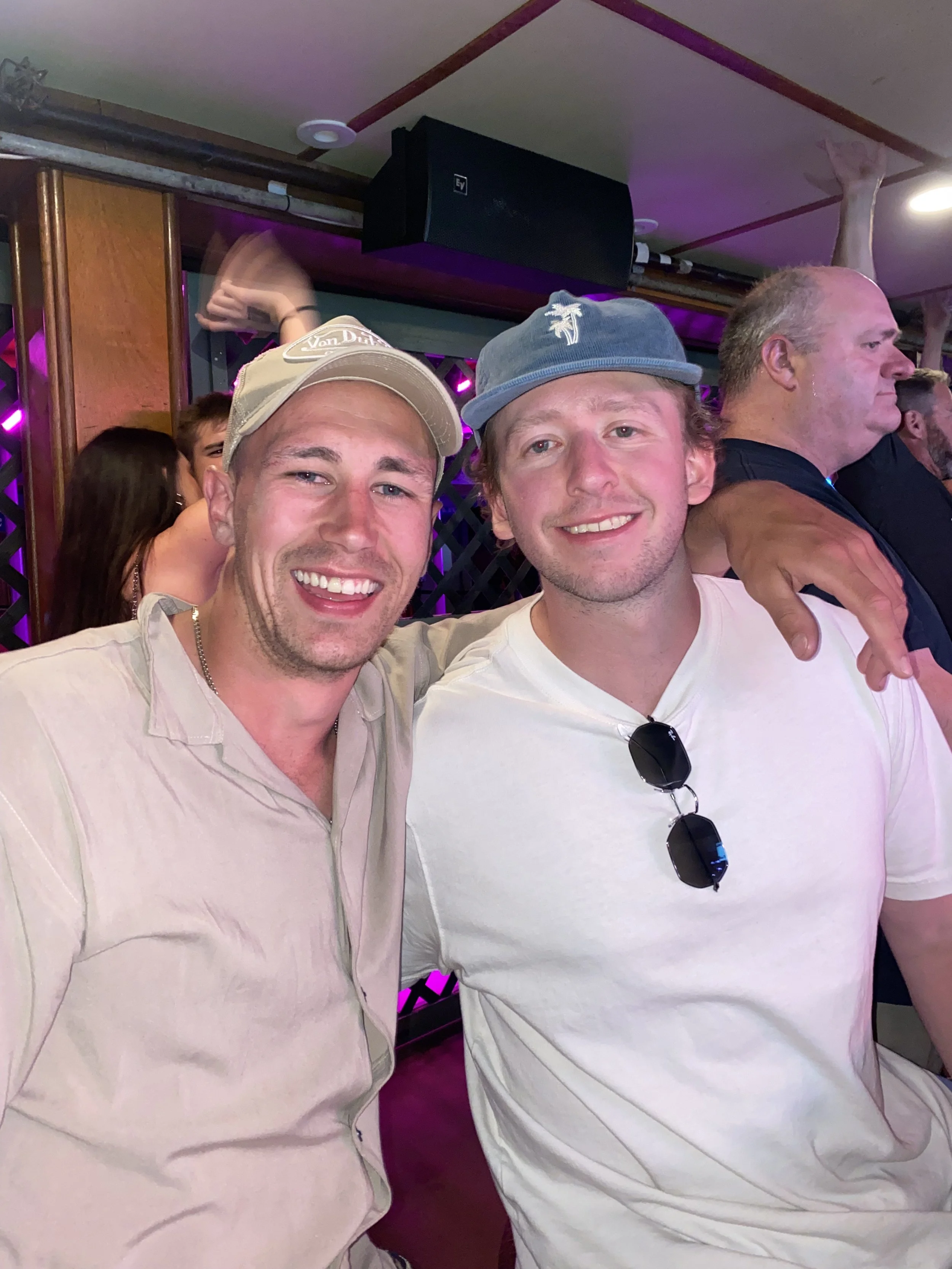 Two young men smiling and posing together at a social event, with one wearing a beige baseball cap and the other wearing a light blue cap and sunglasses hanging on his shirt, in a venue with purple lighting.