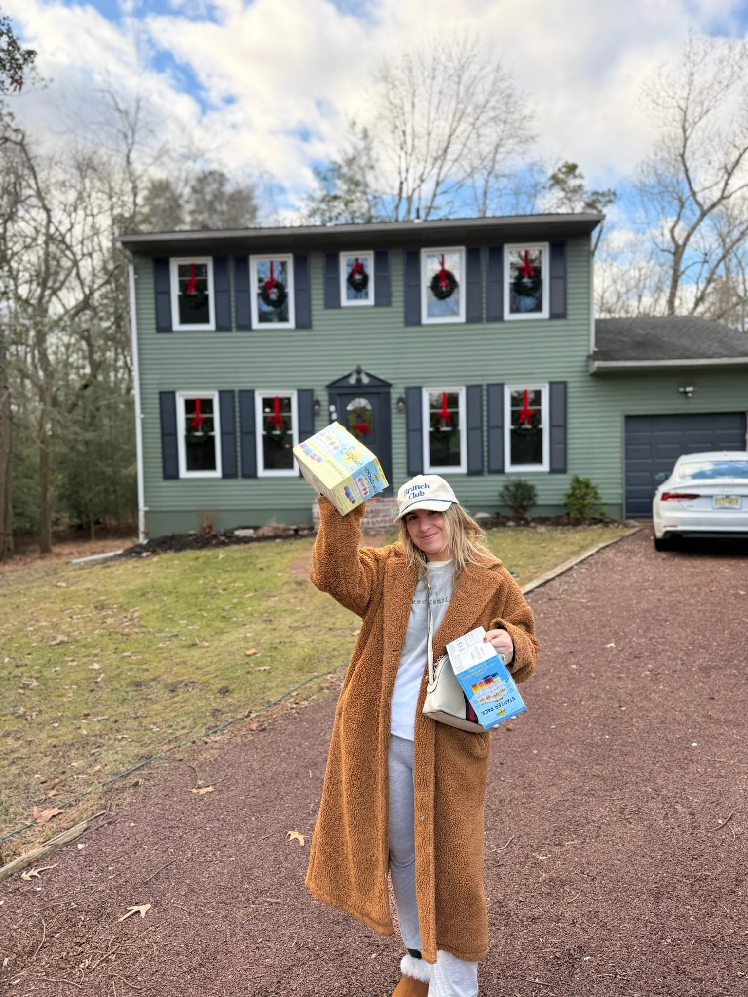 Woman standing on driveway holding grocery items, wearing a brown coat, white cap, grey pants, and white shoes, in front of a green house decorated with Christmas wreaths and red bows in the windows.