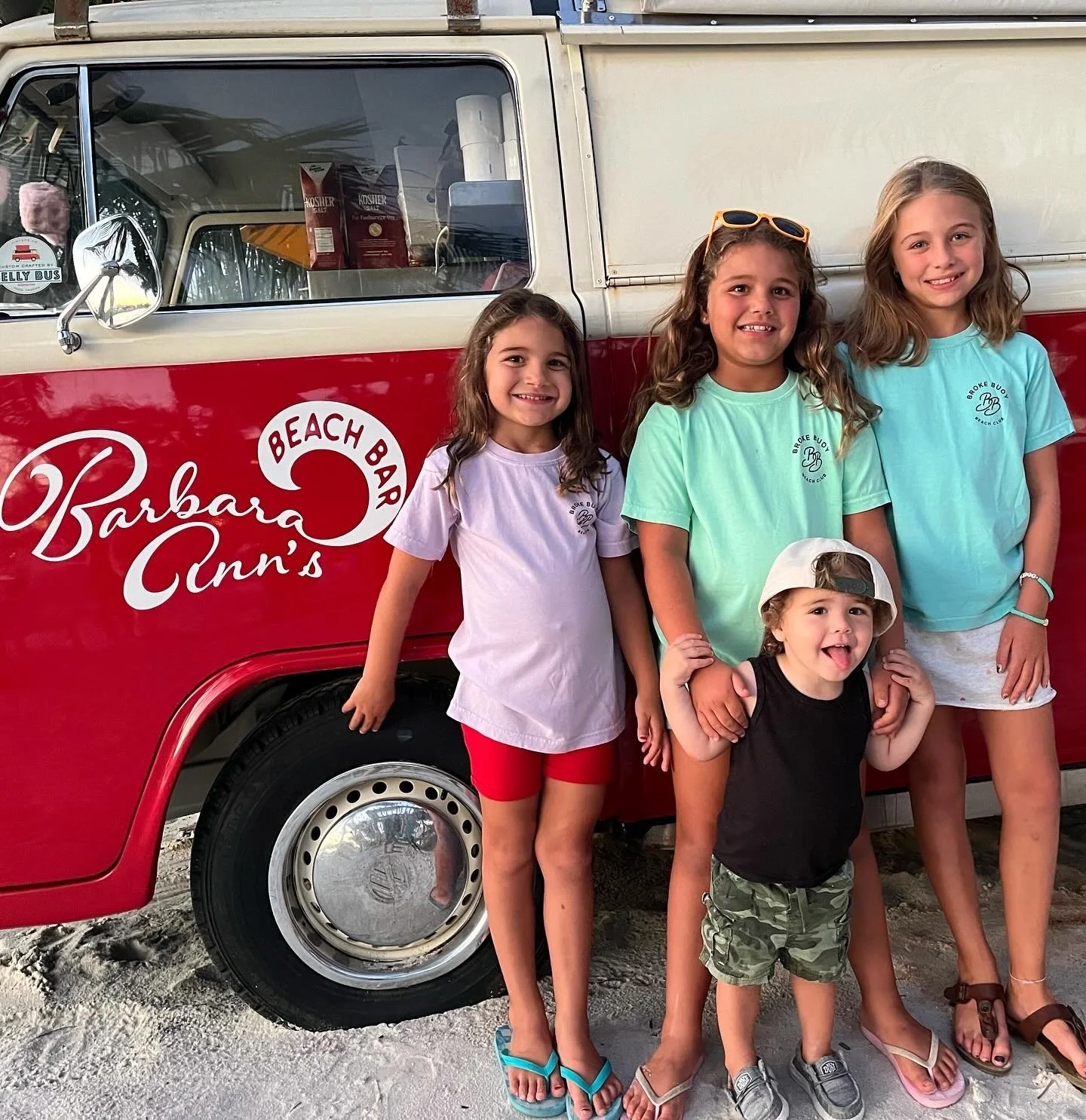 Four girls standing in front of a red and white beach-themed van with the words 'Barbara Ann's Beach Bar' on the side. The girls are smiling, and the youngest is wearing a backward cap and making a funny face.