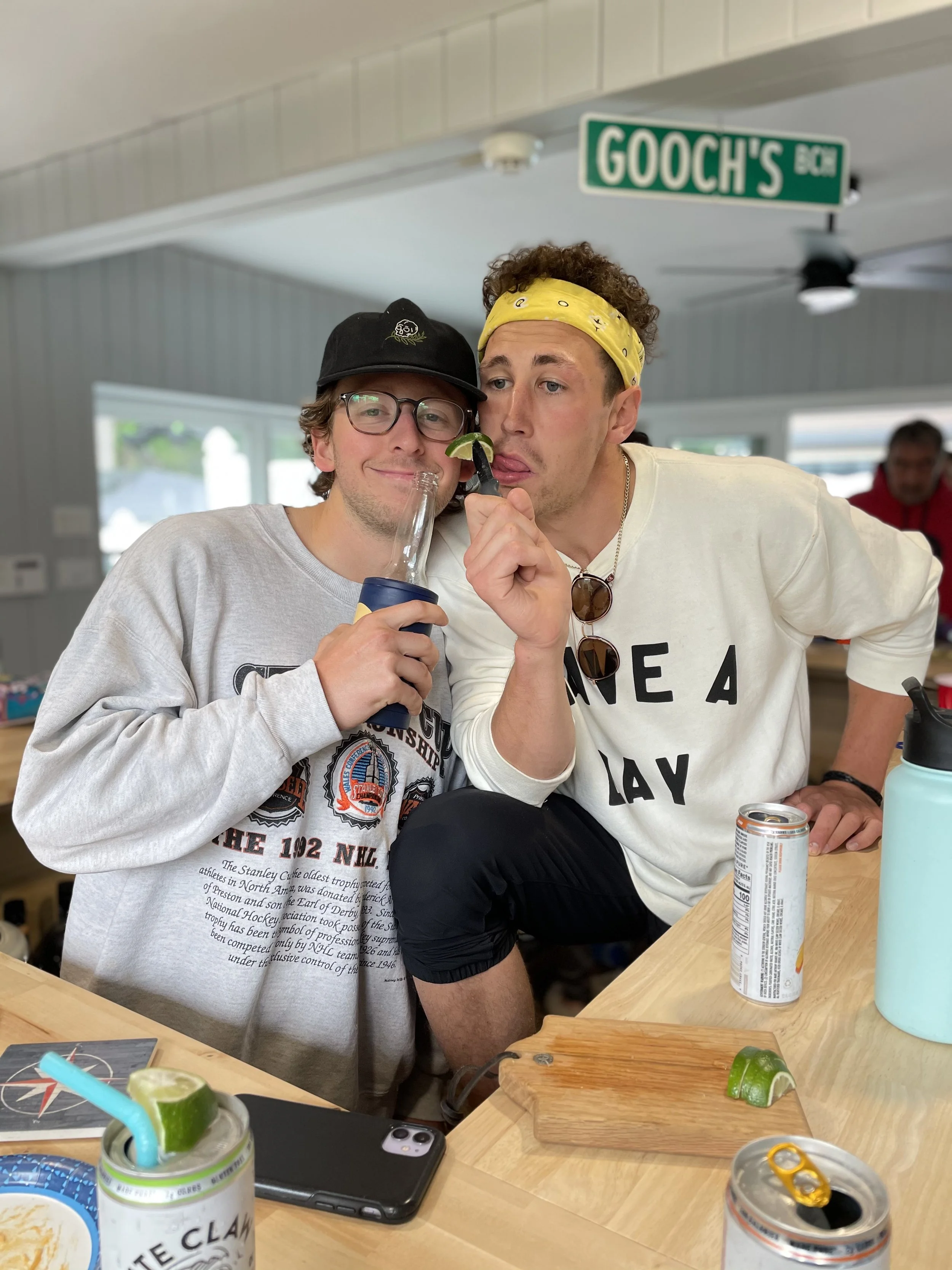 Two young men inside a casual dining area, one holding a bottle and the other holding a slice of lime near their mouth, both appear to be enjoying themselves. The background shows a sign that reads 'Gooch's'.