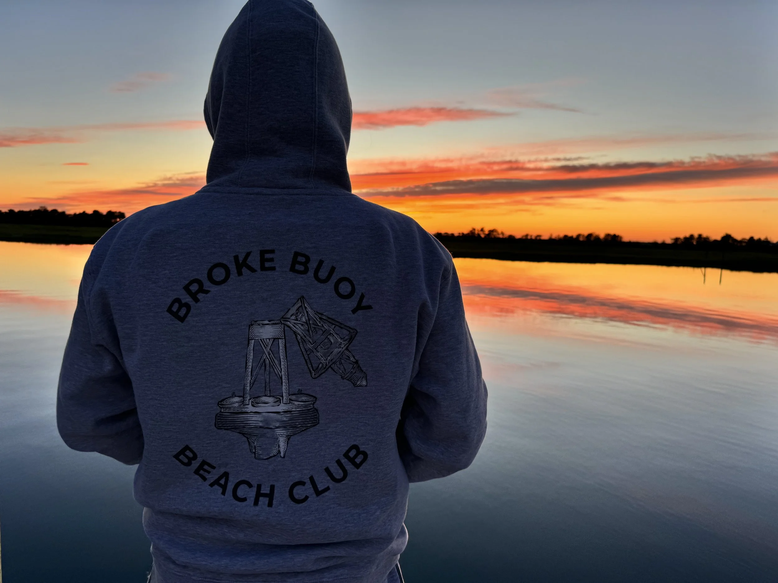 Person wearing a hoodie with 'Broke Buoy Beach Club' logo, standing by a calm body of water at sunset.
