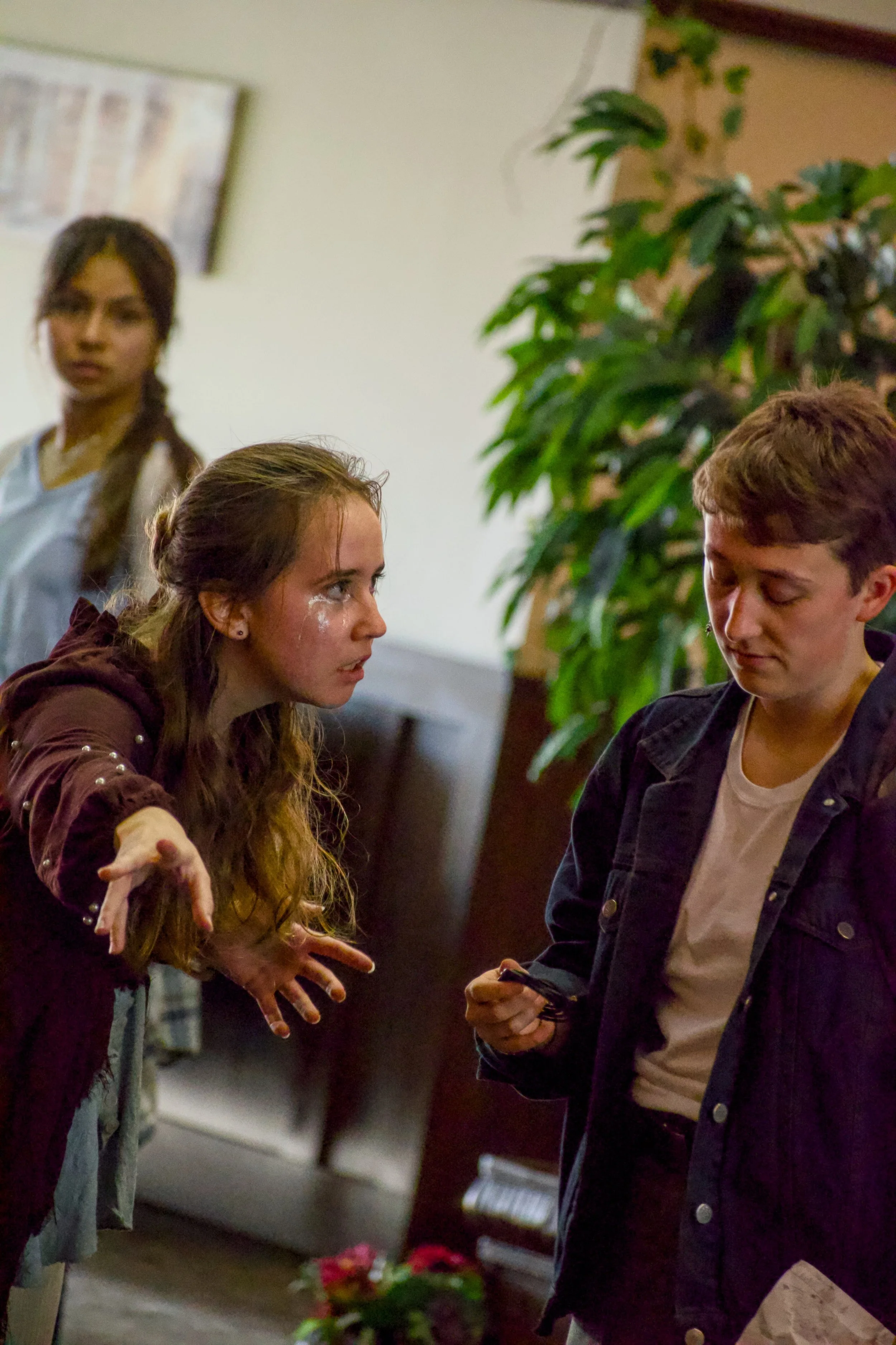 Actor playing an angel in maroon robe watches non-binary person looking at a bracelet, while femme person looks on from the blurry background