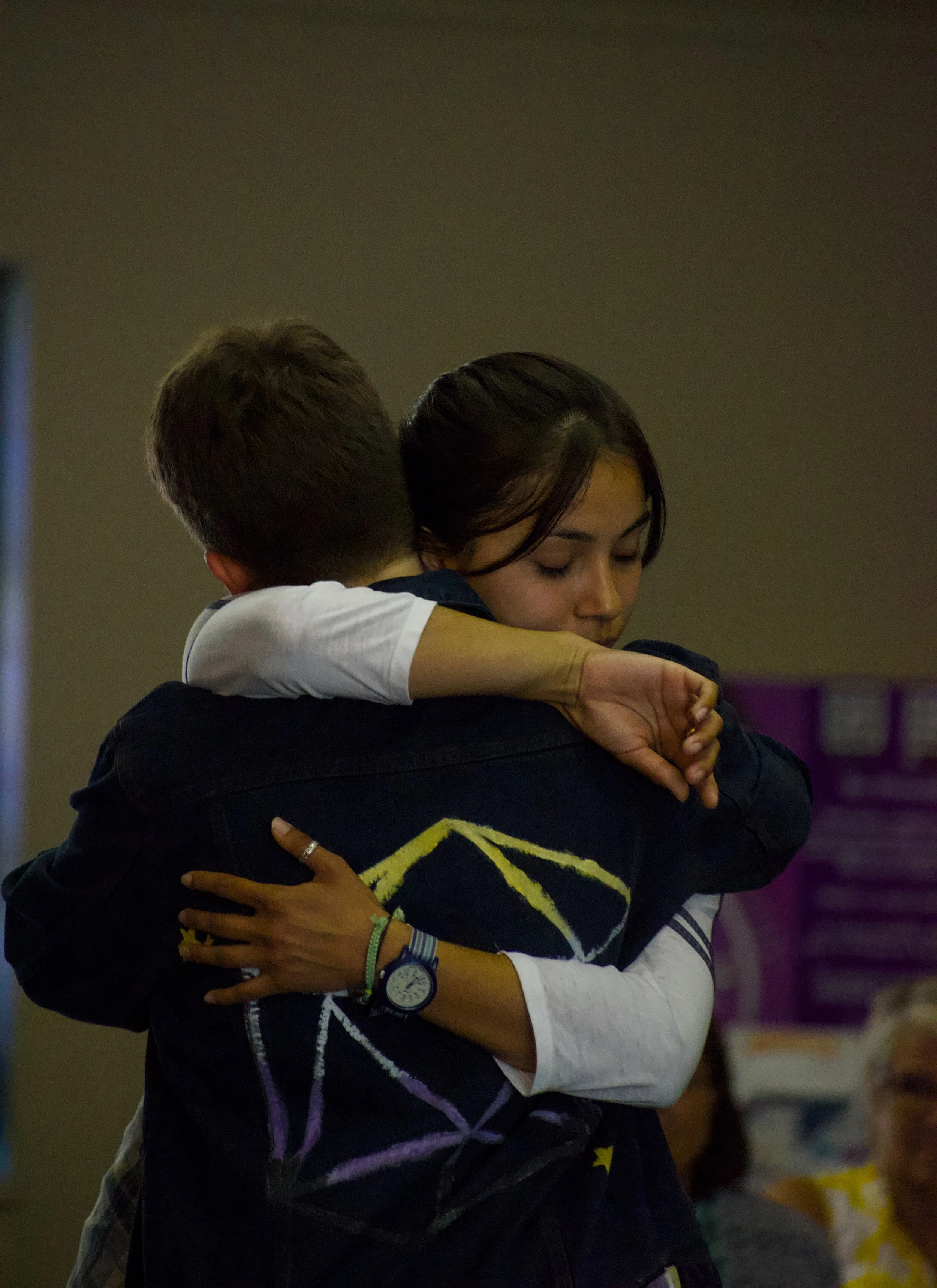 Femme person in white shirt hugs non-binary person in painted denim jacket
