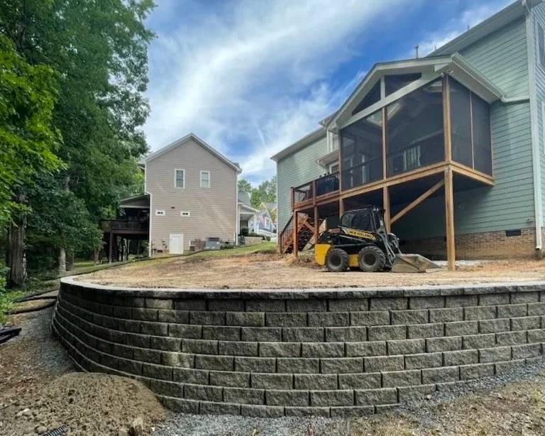Newly built raised patio with retaining wall of interlocking bricks, elevated behind a house with a screened porch, under a partly cloudy sky, surrounded by some trees and other houses.
