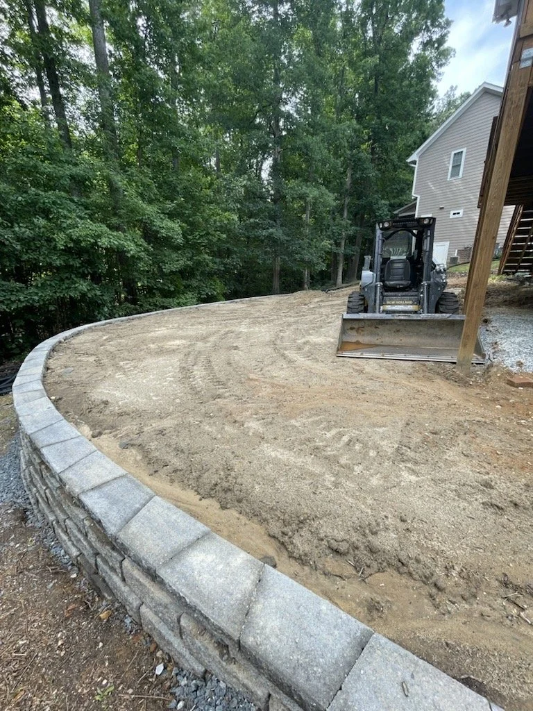 Construction site with a curved stone retaining wall, dirt ground, a small bulldozer, and a house in the background surrounded by trees.