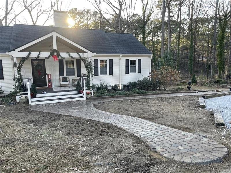 White house with black shutters, decorated for Christmas with a wreath and garland, front porch with two chairs, brick walkway leading to the porch, unpaved yard, neighboring trees.