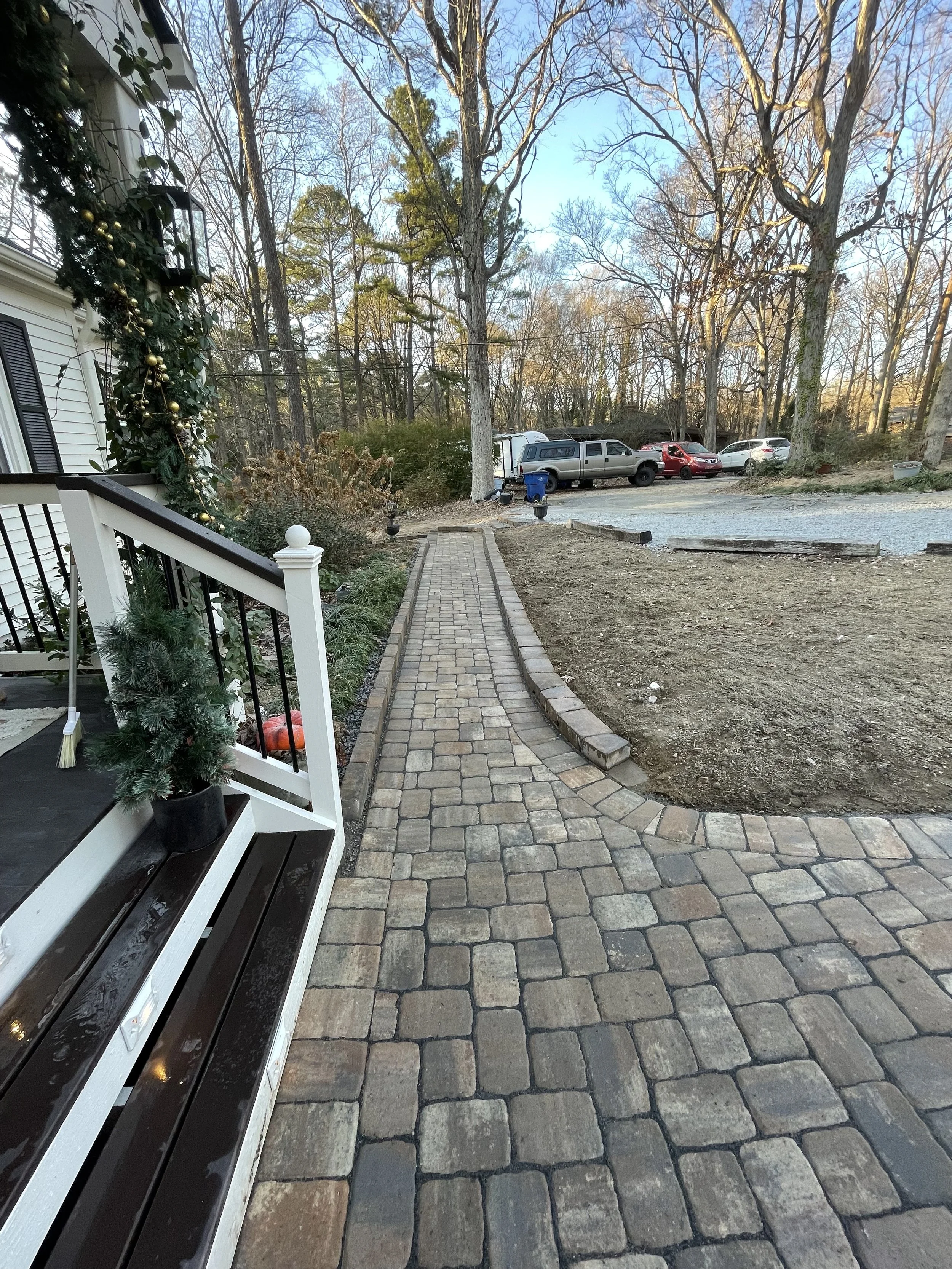 A paved stone walkway leading away from a porch decorated with Christmas ornaments; the walkway is bordered by a garden bed with greenery and a railing on the left, and an area of dirt on the right, with leafless trees and a parking lot with several 