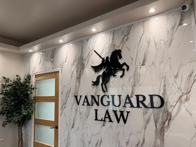 Interior of a law firm's office lobby with a marble wall. The wall features a black silhouette of a knight on a horse, holding a sword, above the words "Vanguard Law" in large black letters. There is a door with frosted glass panels and a potted plant nearby.
