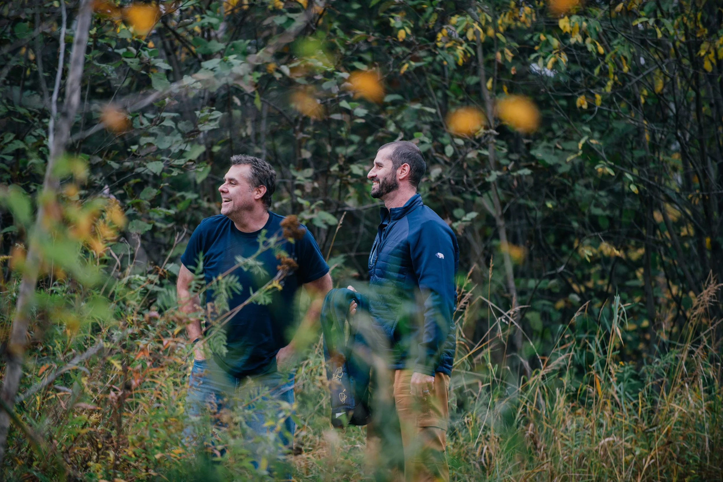 Two men laughing and talking while walking through a dense, green forest with tall grass and leafy trees.