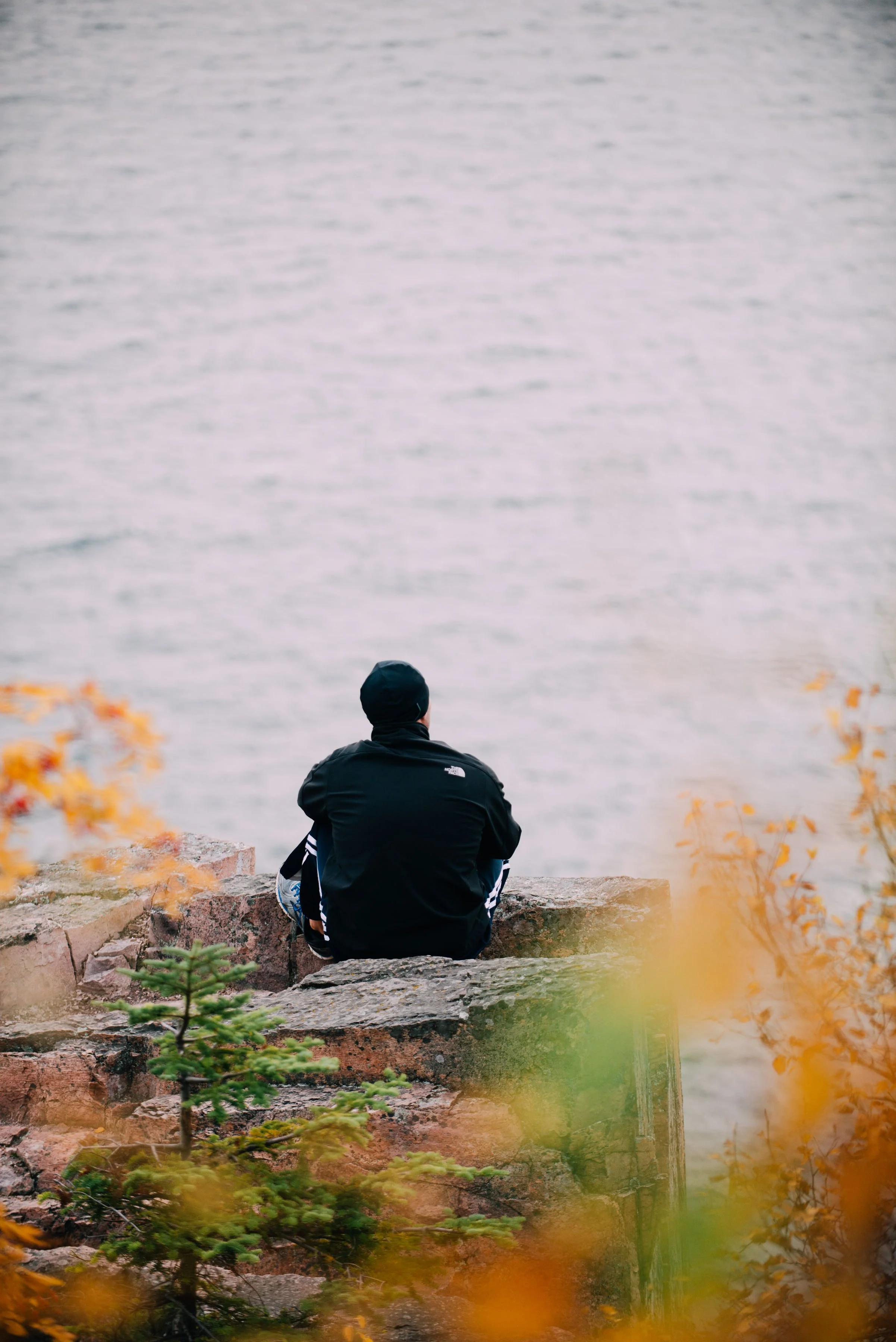 Person sitting on rocks by a body of water, facing away, wearing a black jacket and black cap, surrounded by autumn leaves.