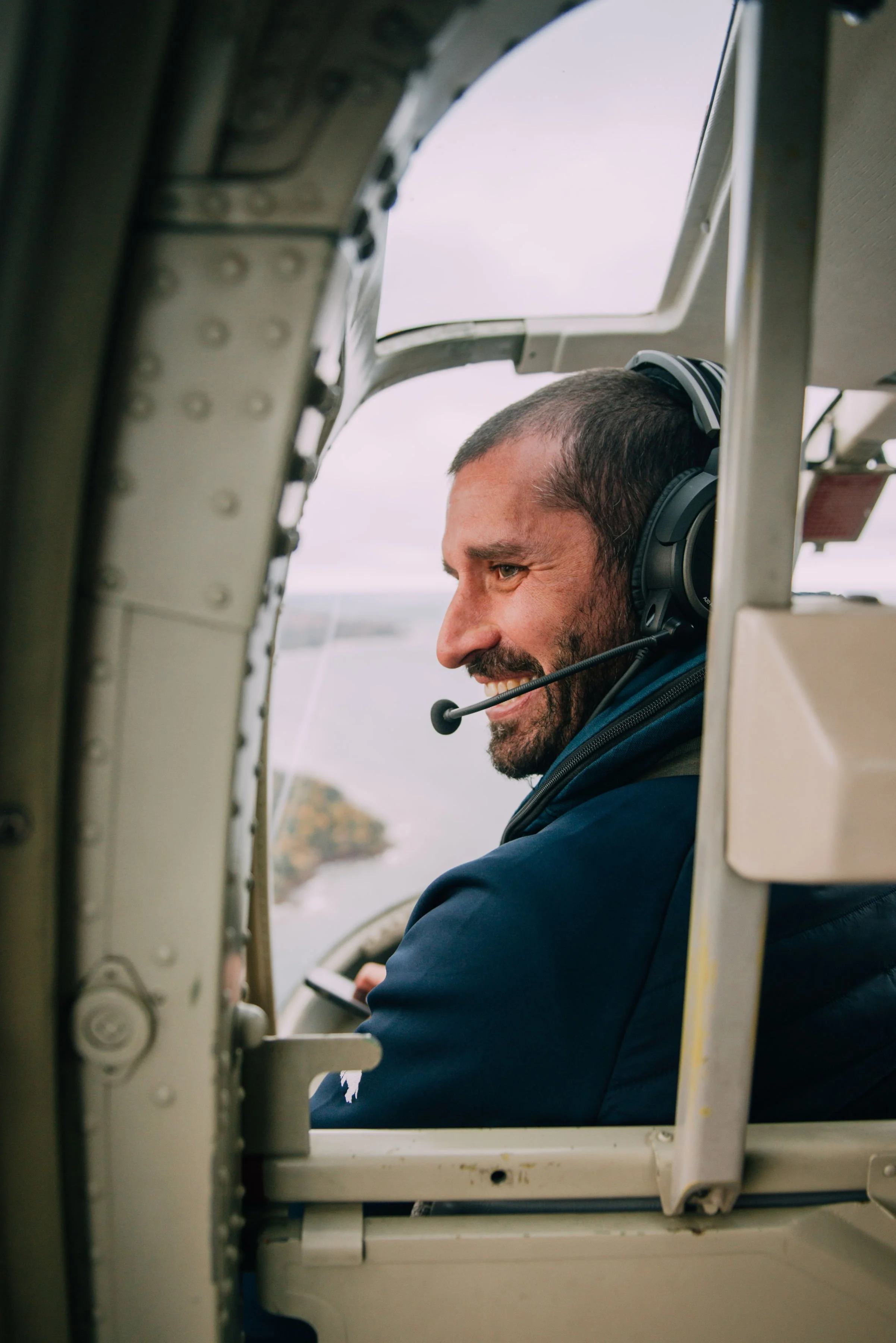 A smiling man with a headset sitting inside a helicopter cockpit, looking out the window at a body of water and land in the distance.