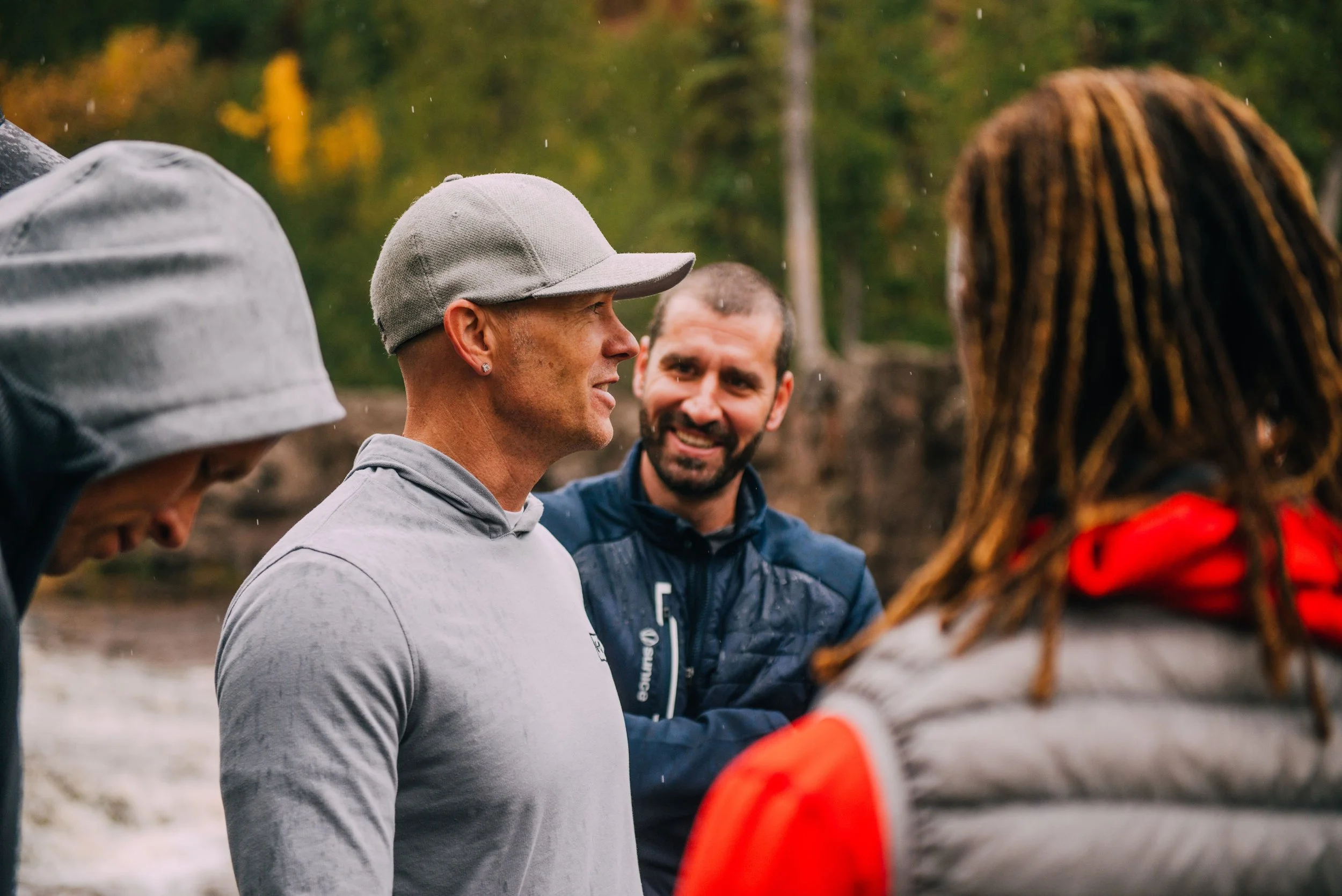 Four people standing outdoors near a river, engaged in conversation, with autumn trees in the background.