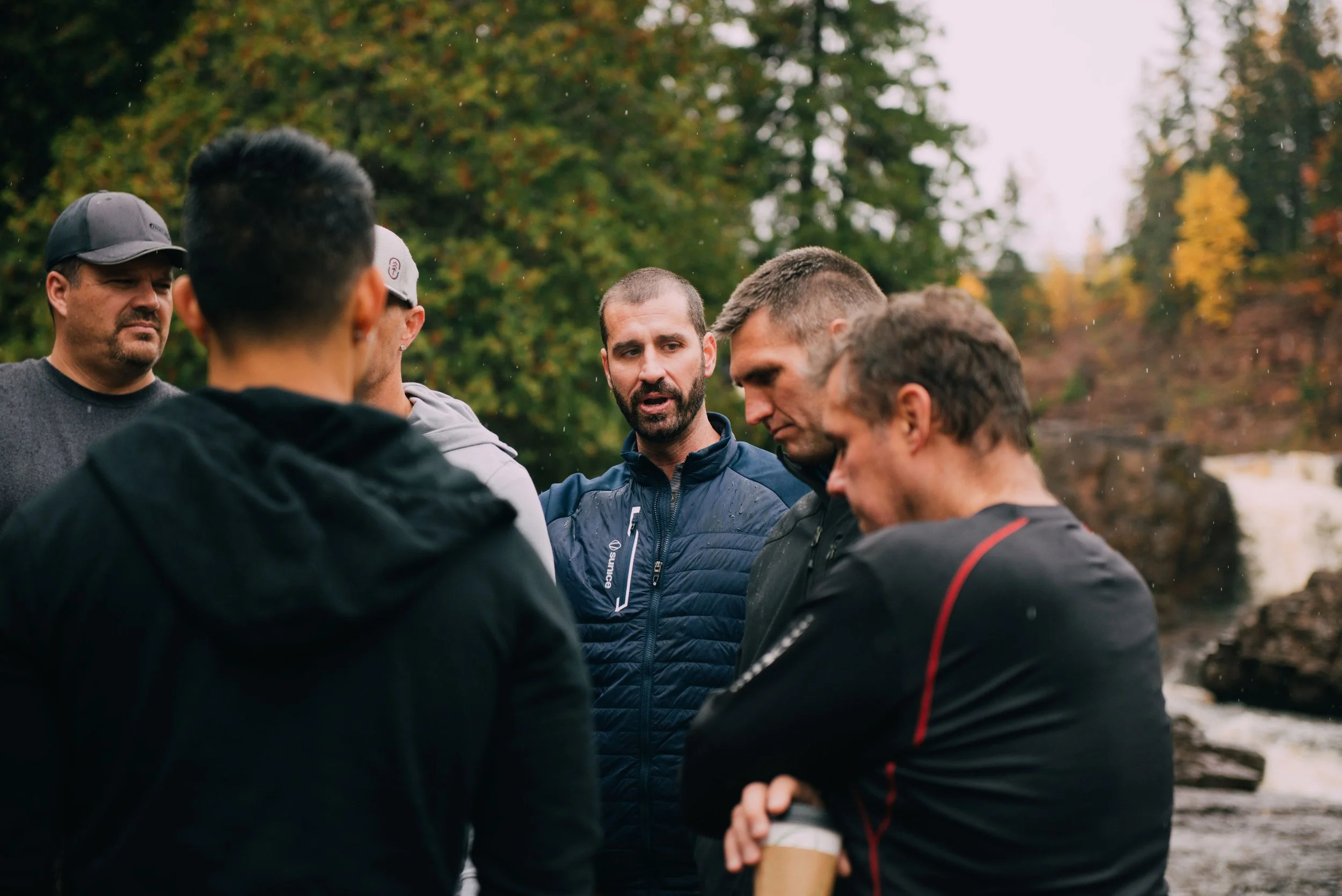 Group of men standing outdoors near a river, engaging in conversation, surrounded by trees with fall foliage.