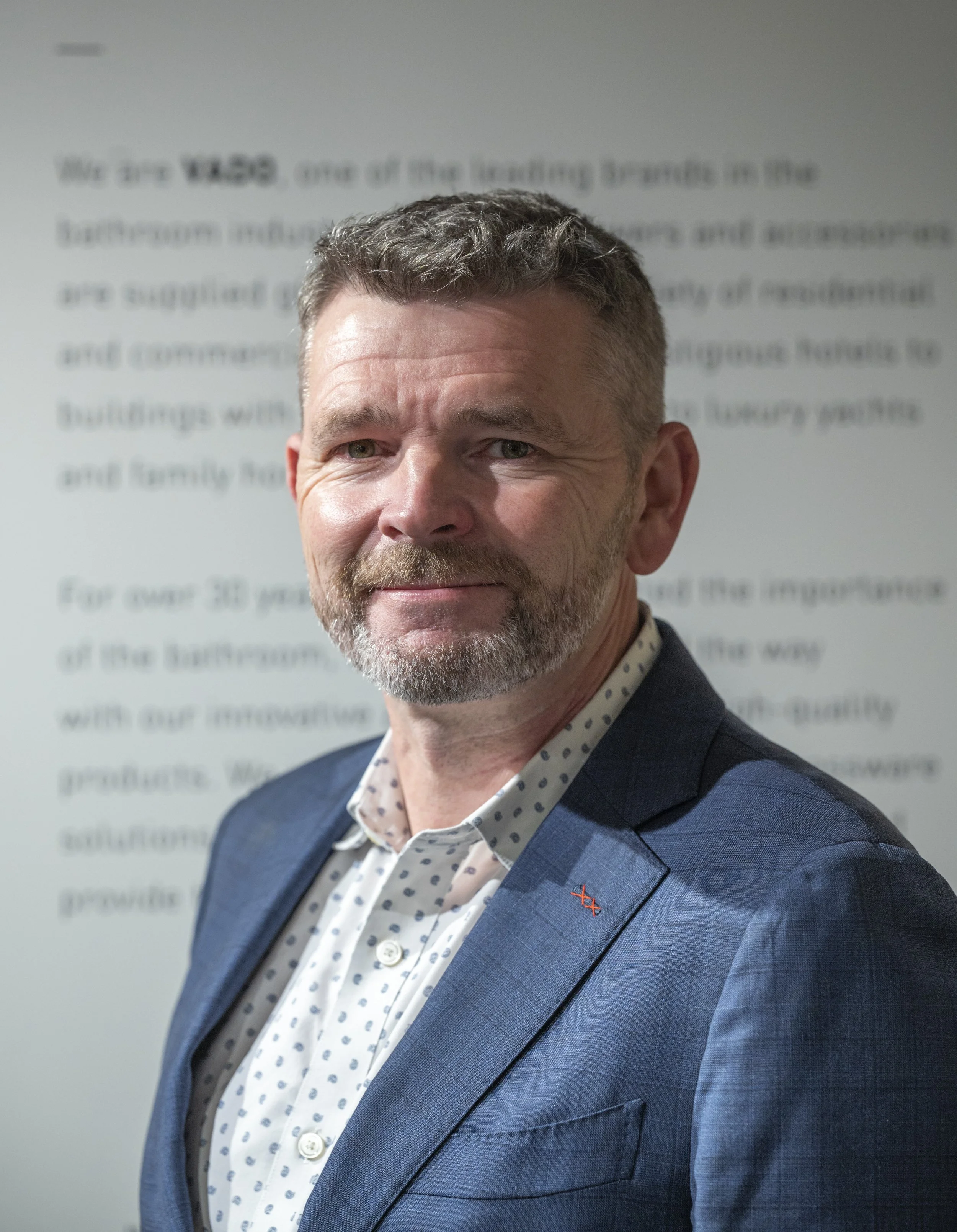 A middle-aged man with short brown hair, a beard, and blue eyes wearing a navy blue suit and a white polka-dot shirt, standing in front of a blurred text background.