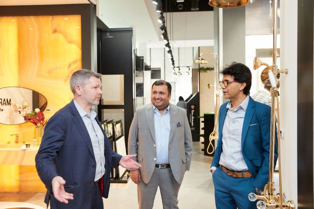 Three men in suits having a conversation in a modern showroom or store, with fixtures of golden faucets and a yellow onyx display in the background.