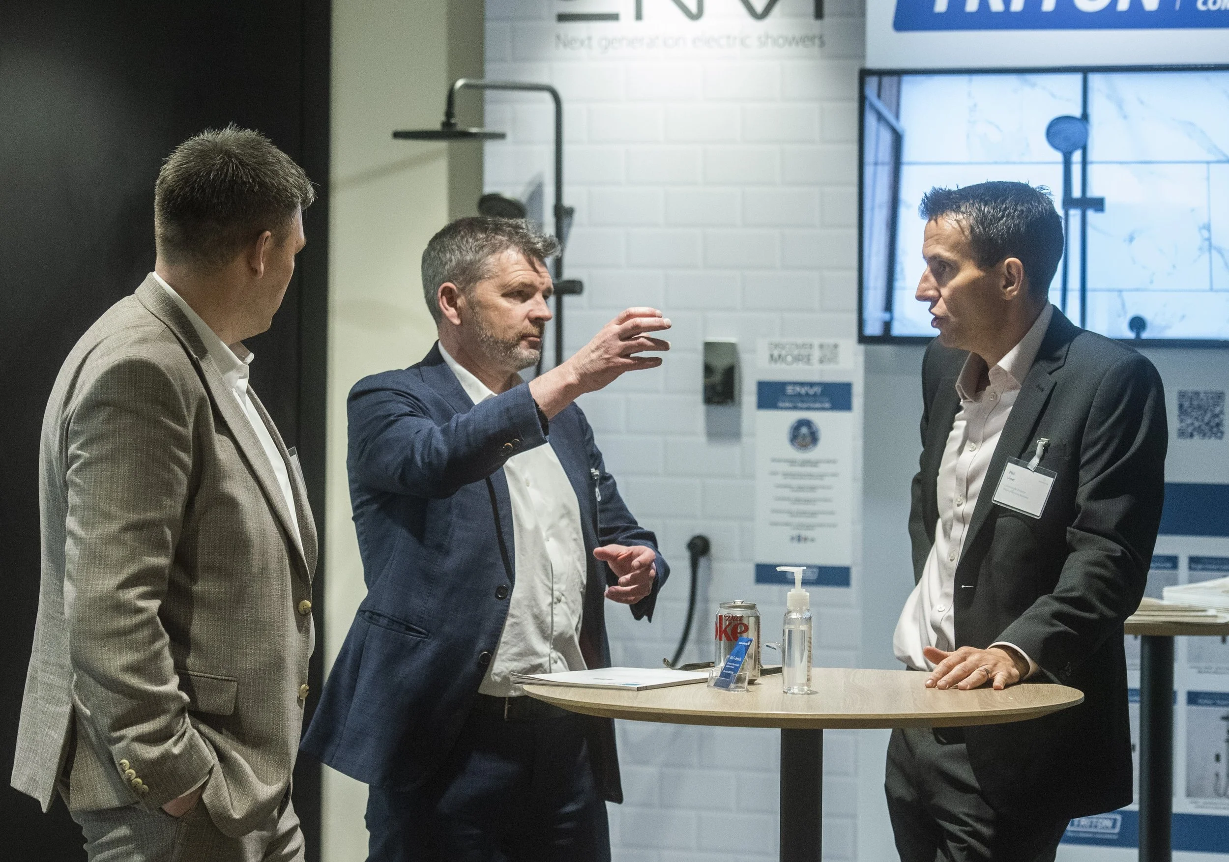 Three men in business attire engaged in conversation at a standing table in a modern setting, with a large screen behind them displaying a technical diagram or presentation.