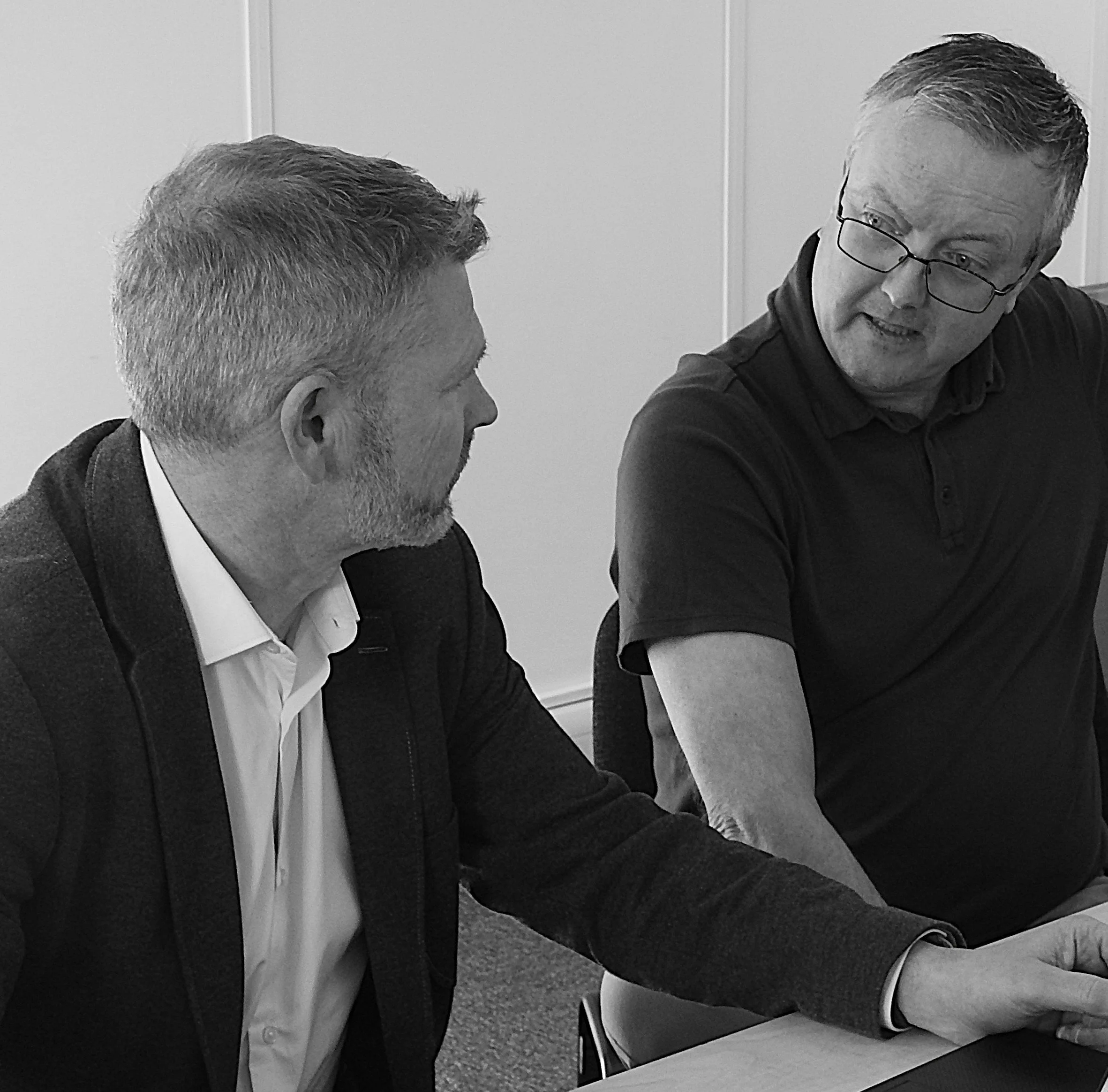 A man giving a presentation in a modern office space with exposed brick walls. He stands next to a large screen displaying the text "Proof ALL HANDS 4/24/2019". An audience of young adults is seated, listening attentively. The office has large windows, desks, computers, and decorative string lights.