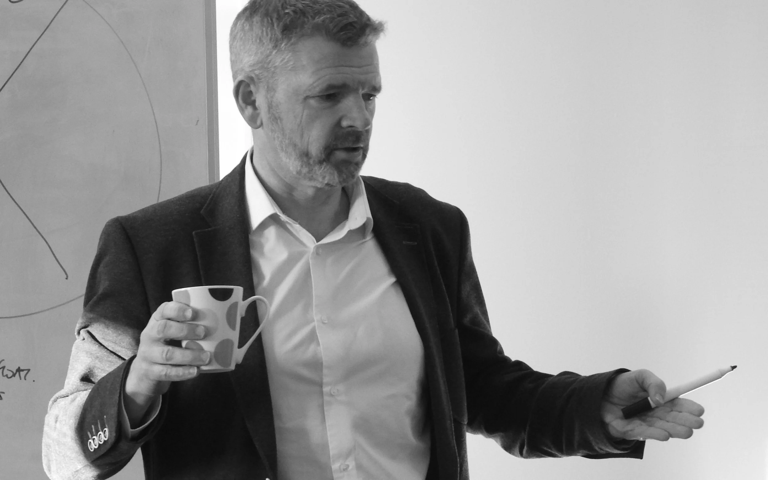 A person pointing at notes and diagrams on a wall, with reflection of the person and notes on a shiny surface, in a room with blinds and overhead shelf.