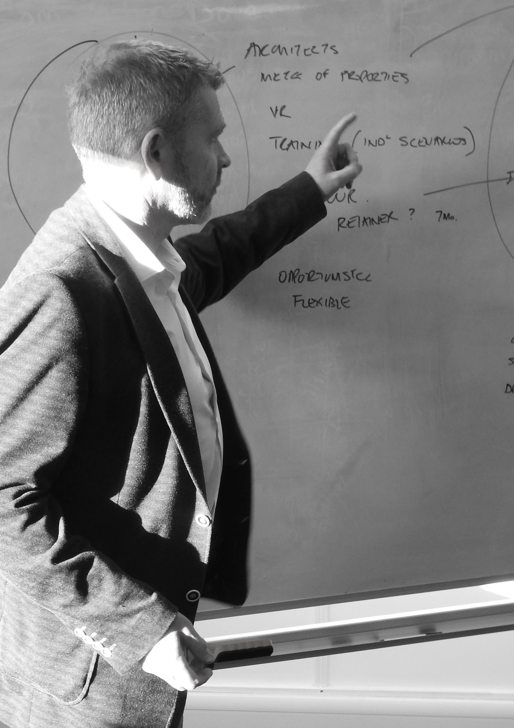 A person giving a presentation to a small group of people in a modern office space with exposed brick walls and large windows. A screen displays the text 'Proof ALL HANDS 4/24/2019'.