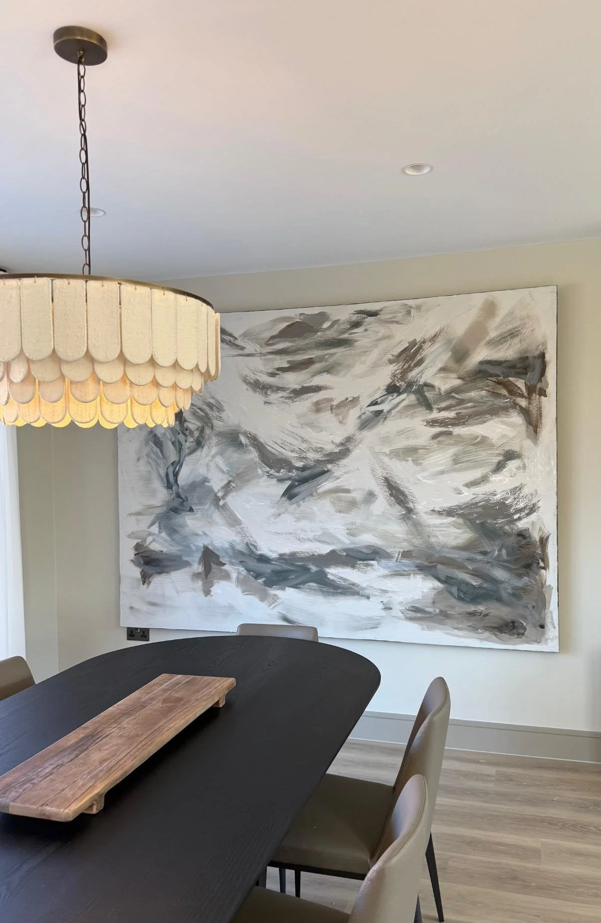 A dining room with a black table, beige chairs, a large abstract black and white painting on the wall, a hanging lamp with a scalloped beige shade, and a wooden serving board on the table.