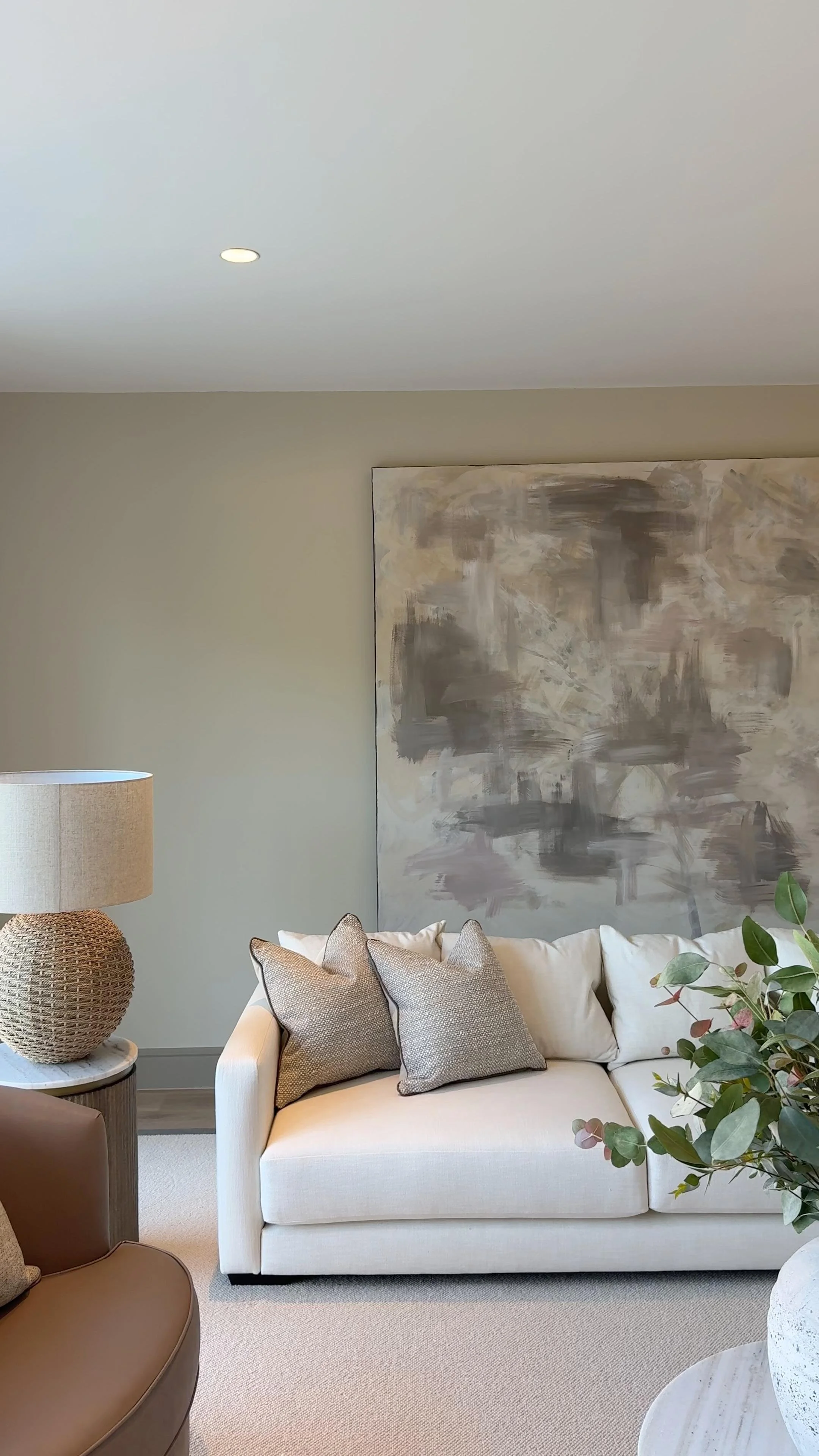 A living room with a white couch, assorted pillows, a side table with a woven lamp, a large abstract painting on the wall, and a plant with green leaves.