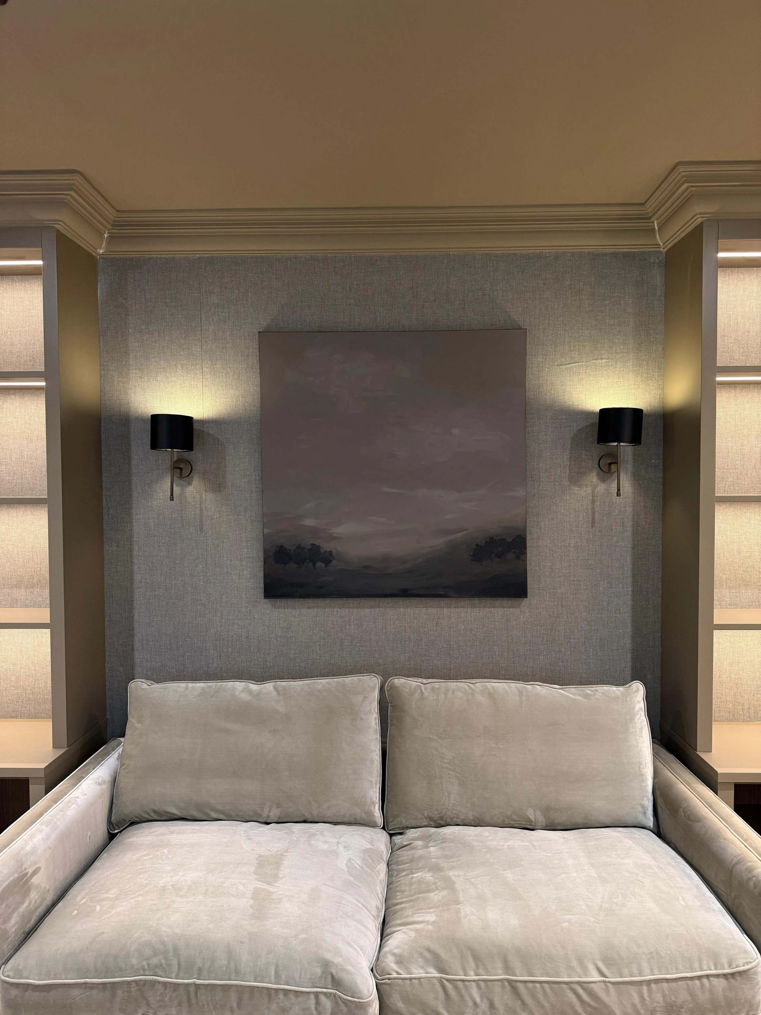A beige sofa with two cushions in front of a textured grey wall and two wall-mounted black lamps on either side of a large landscape painting.