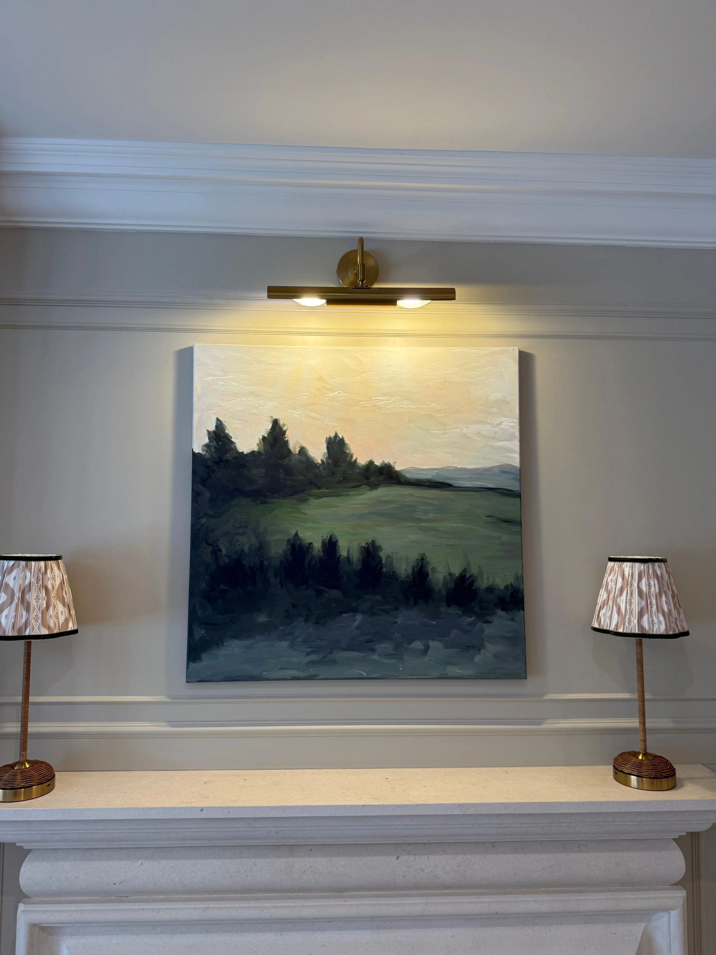 Interior wall with a landscape painting of trees and hills, illuminated by a gold-colored picture light, flanked by two tall floor lamps with patterned lampshades.