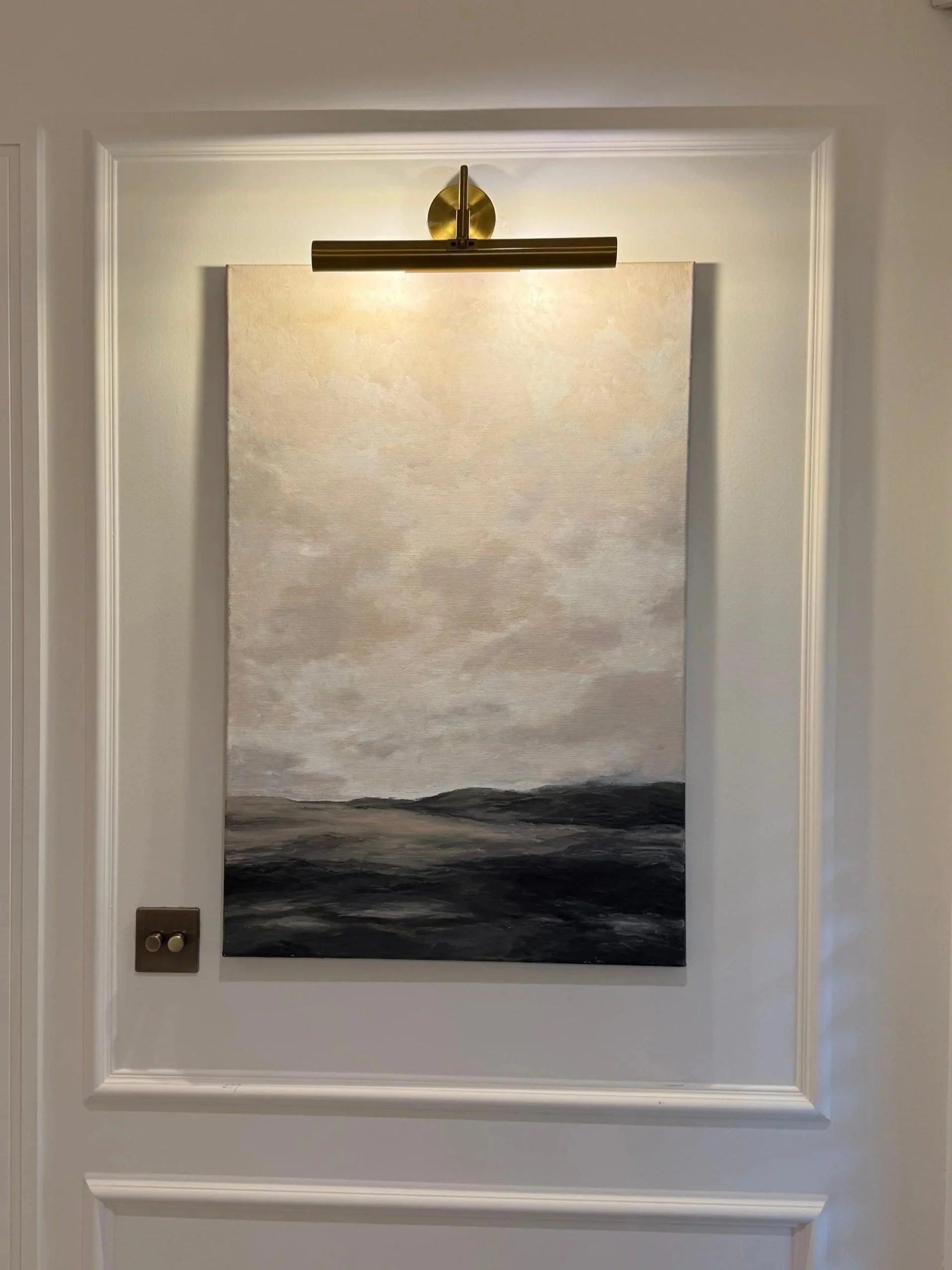A painting of a seascape with dark waves and a cloudy sky, hanging on a white wall above a black floor with white and beige walls surrounding it.