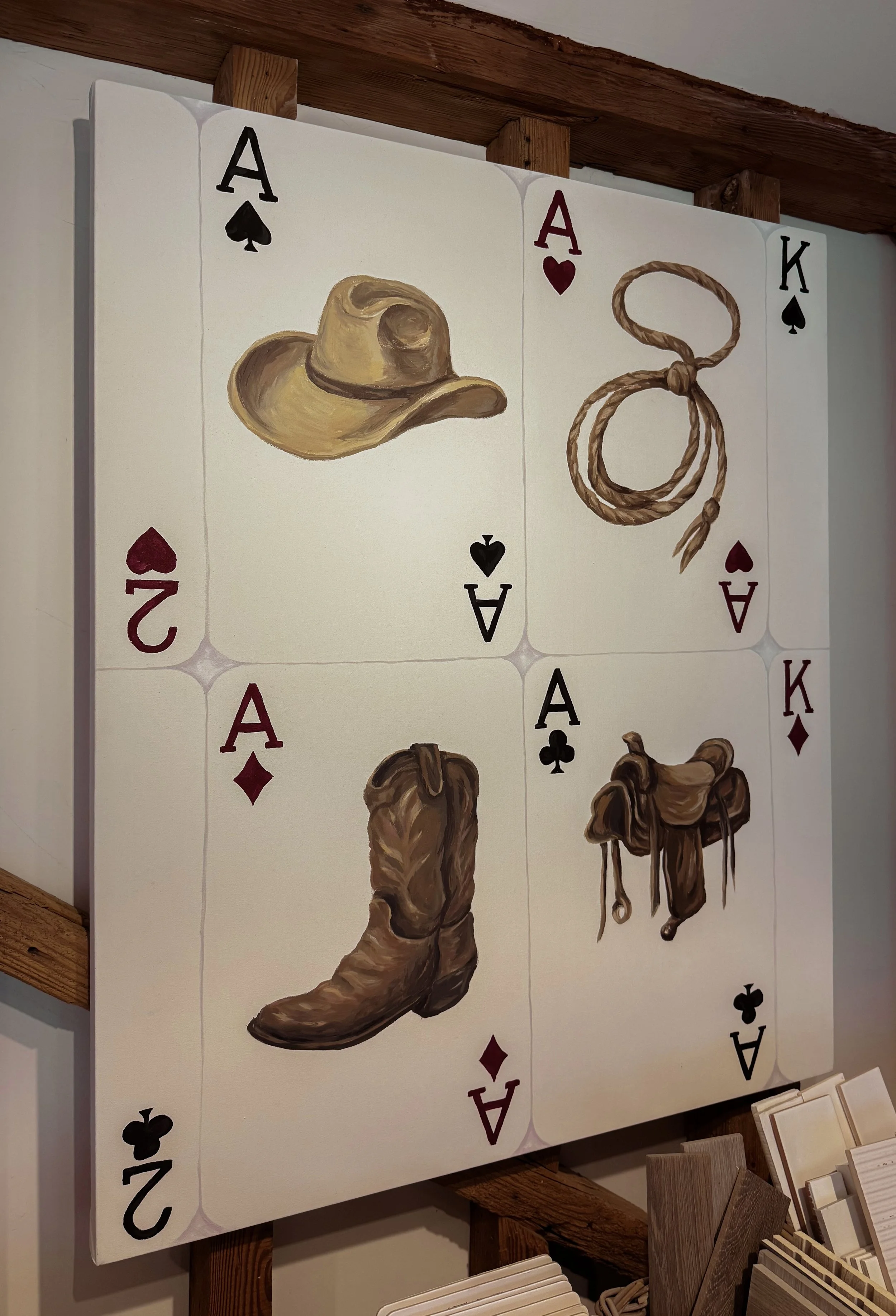 A painting of playing cards with a cowboy hat, a rope, and cowboy boots painted on an acrylic sheet, set against a wooden background.