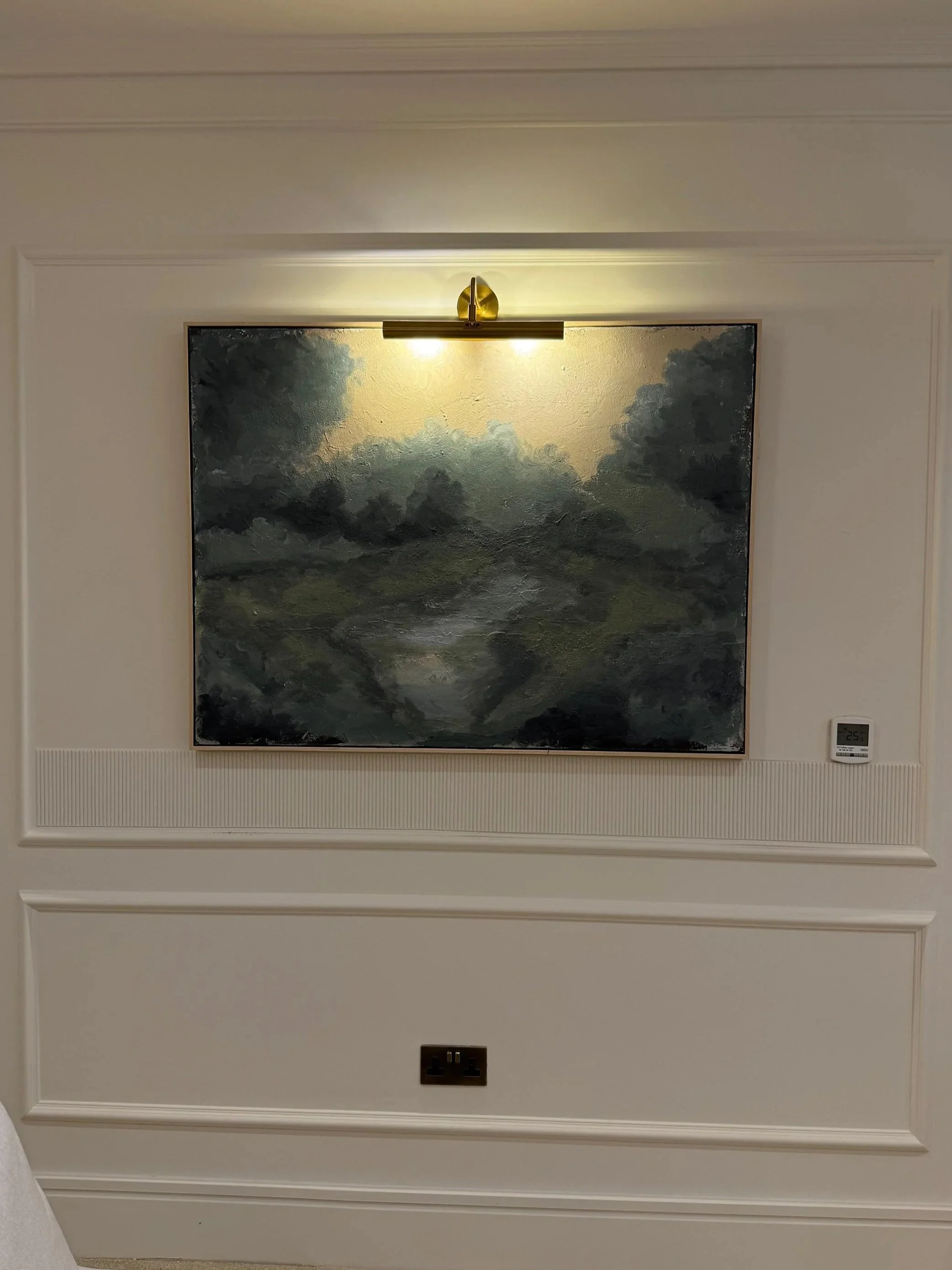 A wall with white molding, a large framed painting of dark stormy clouds, a gold picture light above the painting, a digital thermostat reading 25°C on the right, and a black electrical outlet below the painting.