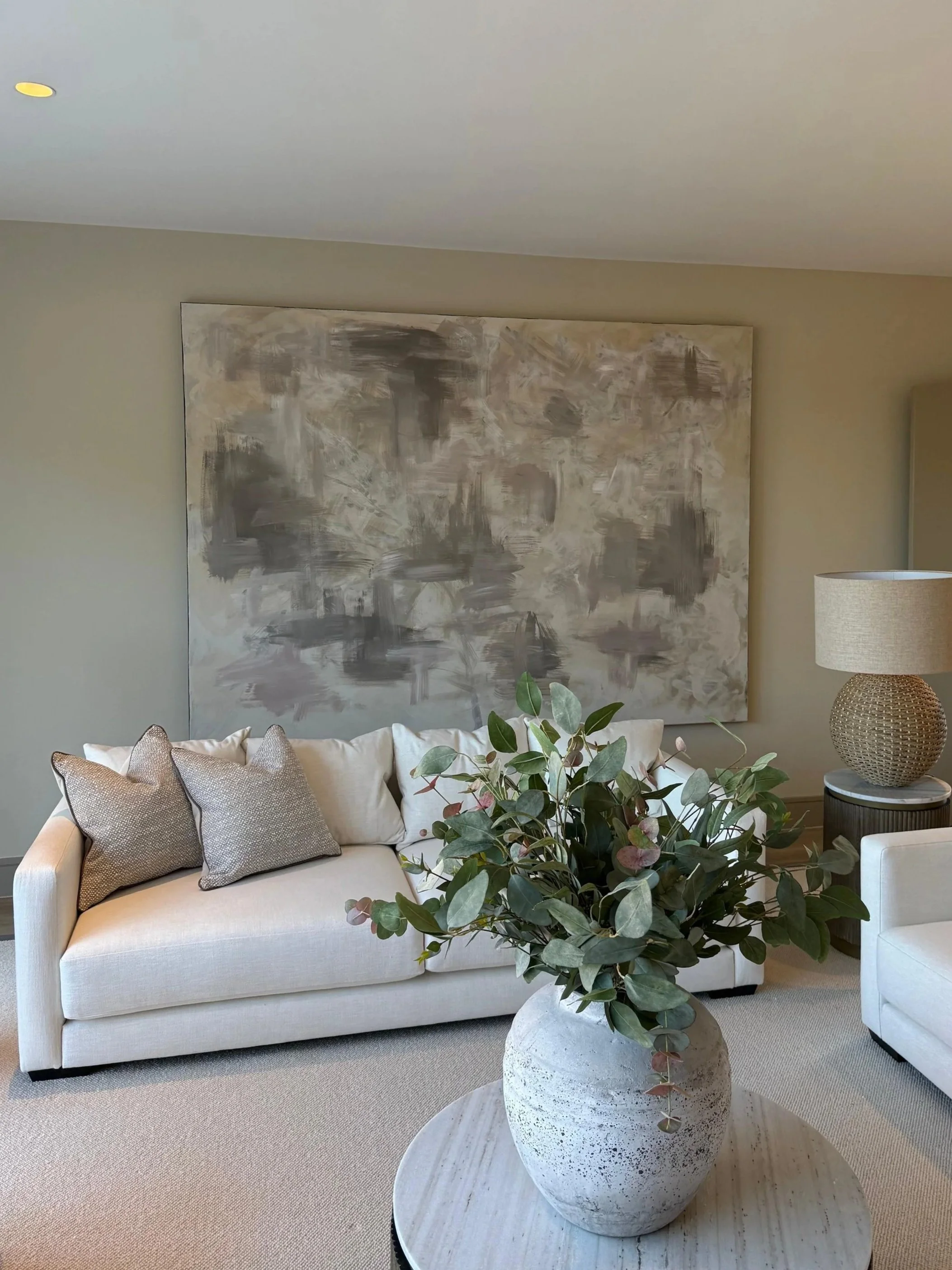 A living room with a white sofa decorated with beige and gray pillows, a large abstract painting on the beige wall, a table with a large potted plant, and a table lamp on a side table to the right.