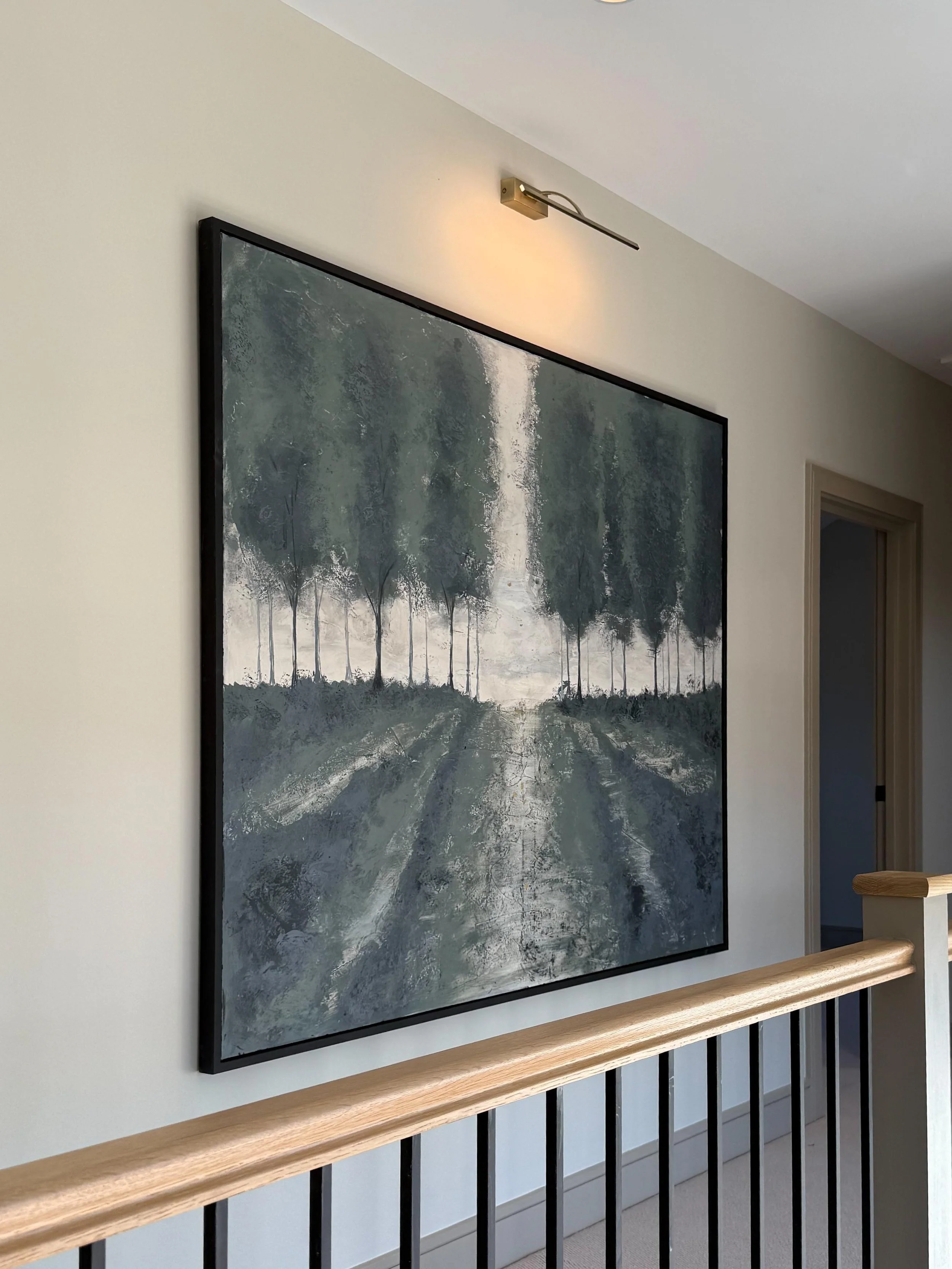 Interior of a modern house showing a large landscape painting of a forest and pathway on the wall, illuminated by an adjustable wall-mounted light fixture.