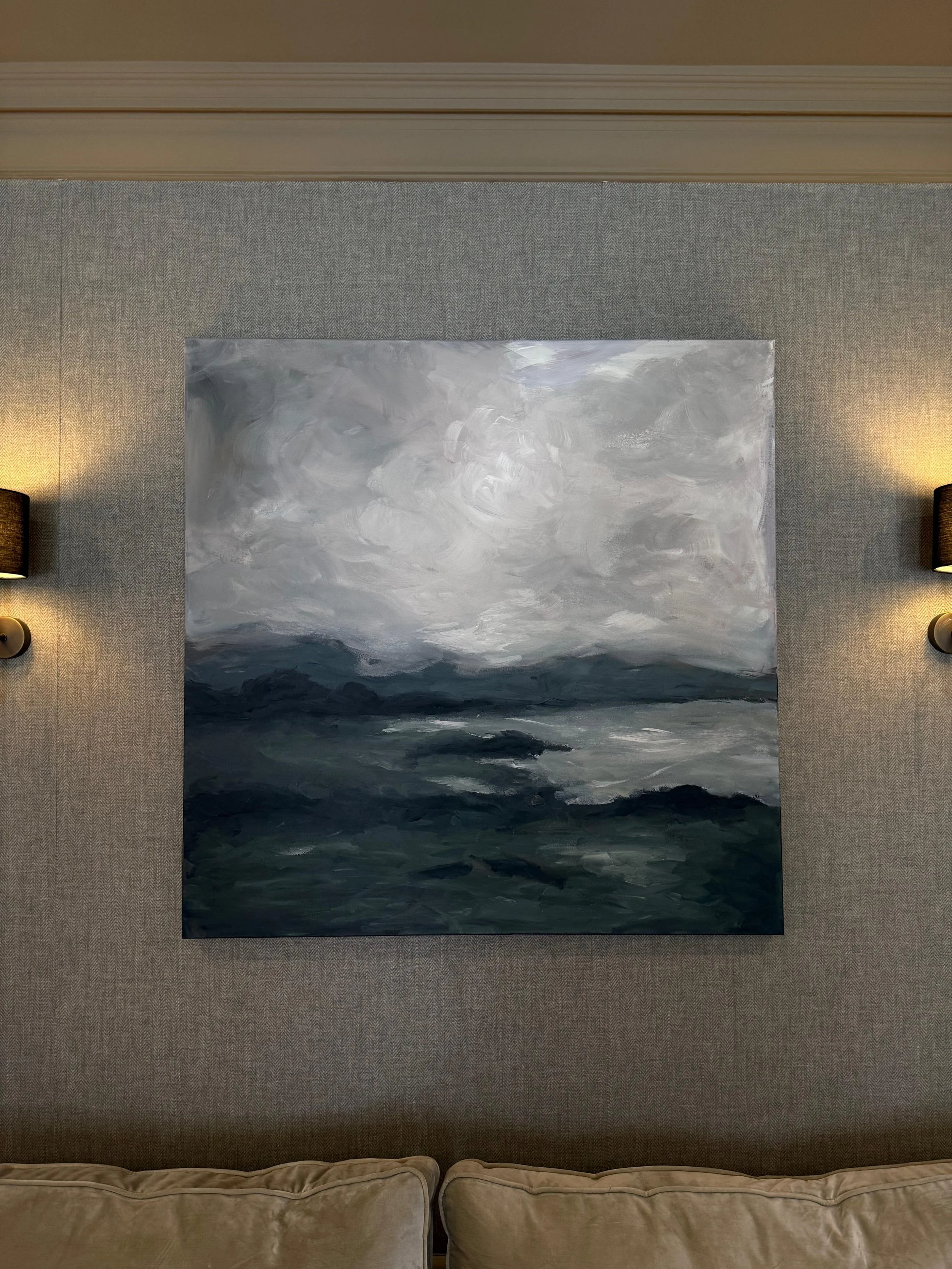 A monochromatic abstract painting with swirling shades of gray and black, hanging on a textured beige wall with two wall-mounted lamps on either side, above a beige sofa.