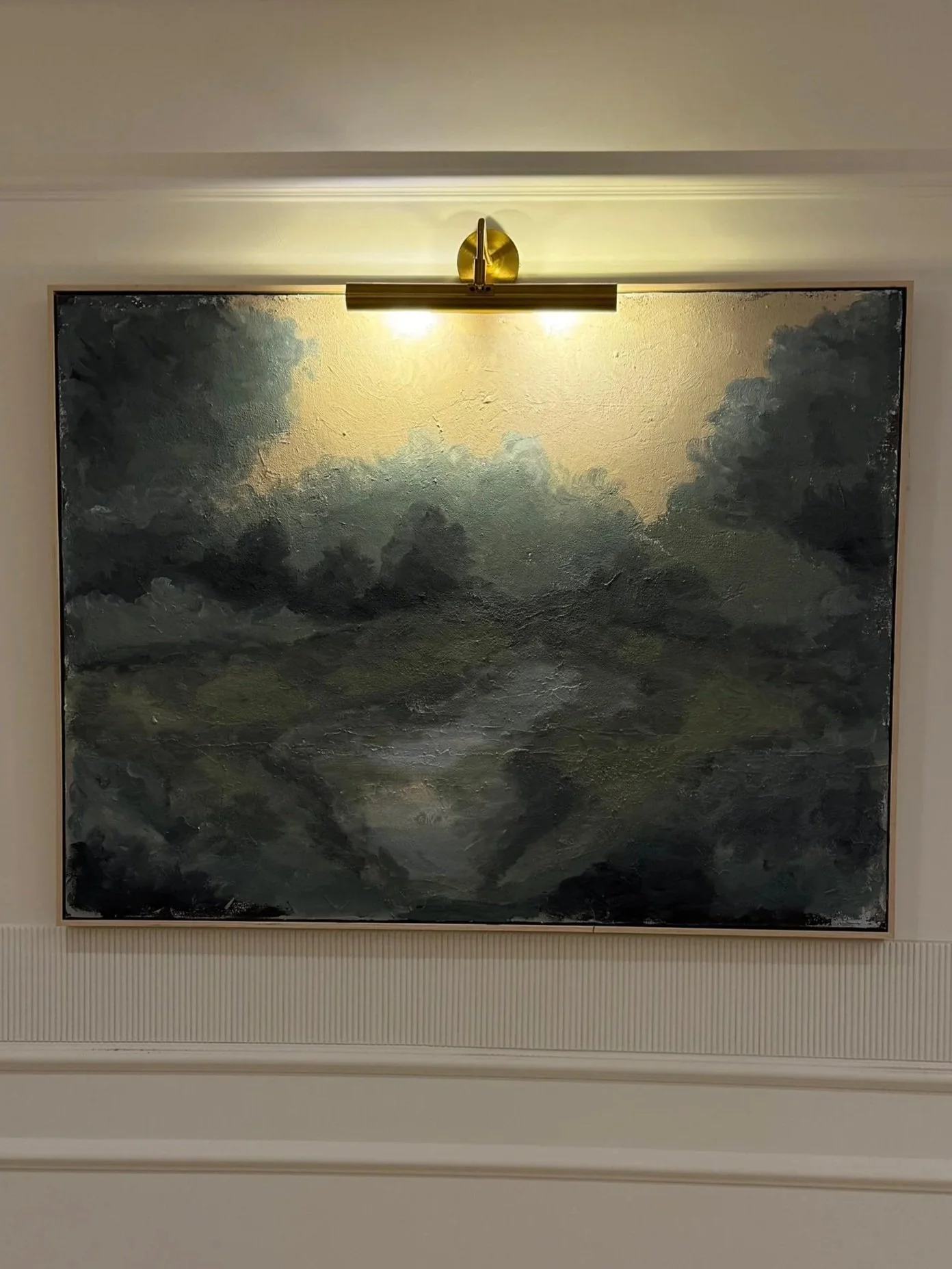 A landscape painting of dark, turbulent clouds over a green landscape, mounted on a wall with a modern brass picture light above it.